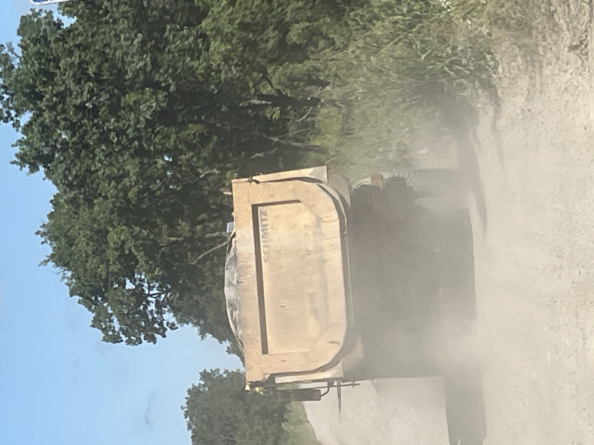 A yellow dump truck is driving down a dirt road, kicking up a cloud of dust. The truck is loaded with sand. The road is lined with trees and vegetation. The sky is blue and there are a few clouds. The scene appears to be in a rural area, possibly outside of Medovo, Bulgaria.