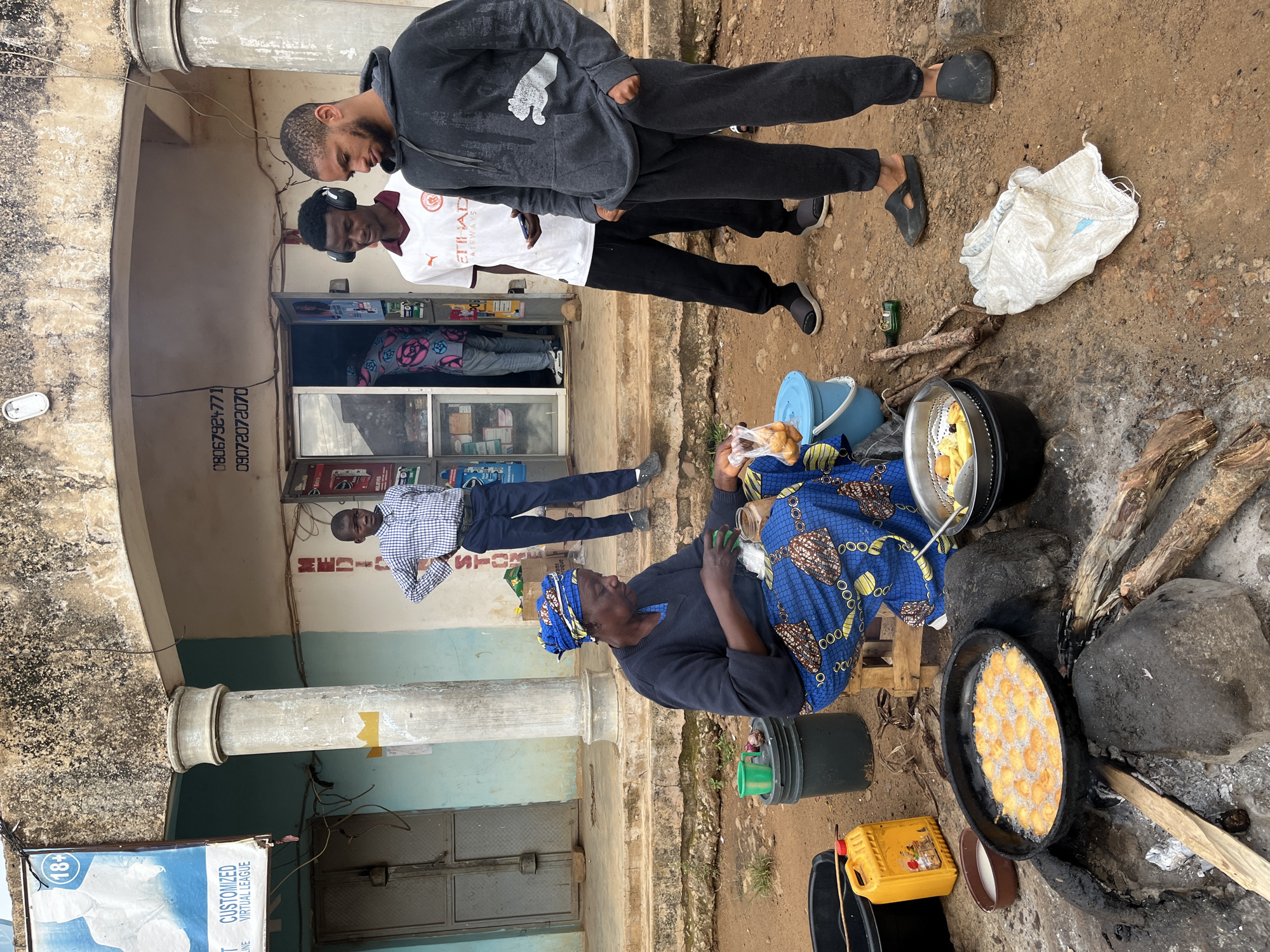 In Jos, Nigeria, a street vendor is frying dough balls in a large pan over an open fire in front of a shop. The vendor, a woman wearing a blue and gold patterned headwrap and dress, is seated on a wooden crate and is in the process of adding more dough to the frying pan. A blue bucket, a green bucket, and a yellow jerrycan are visible near her.
Several young men stand nearby, observing the scene. One young man in a gray hoodie and sweatpants stands with his hands in his pockets, looking down. Another young man in a white shirt with "Etihad Airways" printed on it wears headphones and holds a phone. A third young man, dressed in a checkered shirt and dark pants, stands with his hands on his hips, looking towards the vendor.
The shop behind them appears to be a small retail establishment, with goods visible through its open doorway. The ground is unpaved and dusty, characteristic of an outdoor market or street scene in Jos. A concrete pillar and the weathered facade of the building provide context for the urban environment.