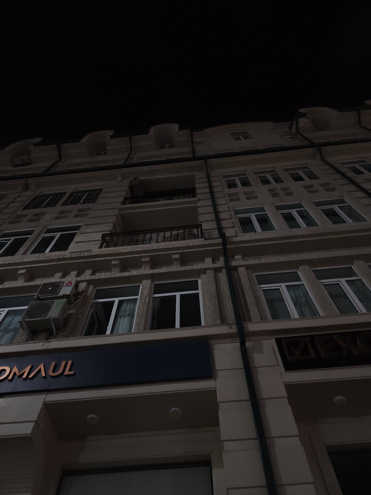 The image shows a low-angle, night-time shot of a multi-story building facade in Fergana City, Uzbekistan. The building appears to be made of light-colored stone with numerous windows and decorative architectural elements. On the lower level, a dark blue signage with bronze-colored lettering is visible, displaying the text "DMAUL". To the right of this signage, another sign with bronze lettering in a square frame is present, with partially visible text that includes "MEX". Two air conditioning units are mounted on the wall near the "DMAUL" sign. The sky above the building is completely dark. The building's design suggests it could be a commercial or residential complex.