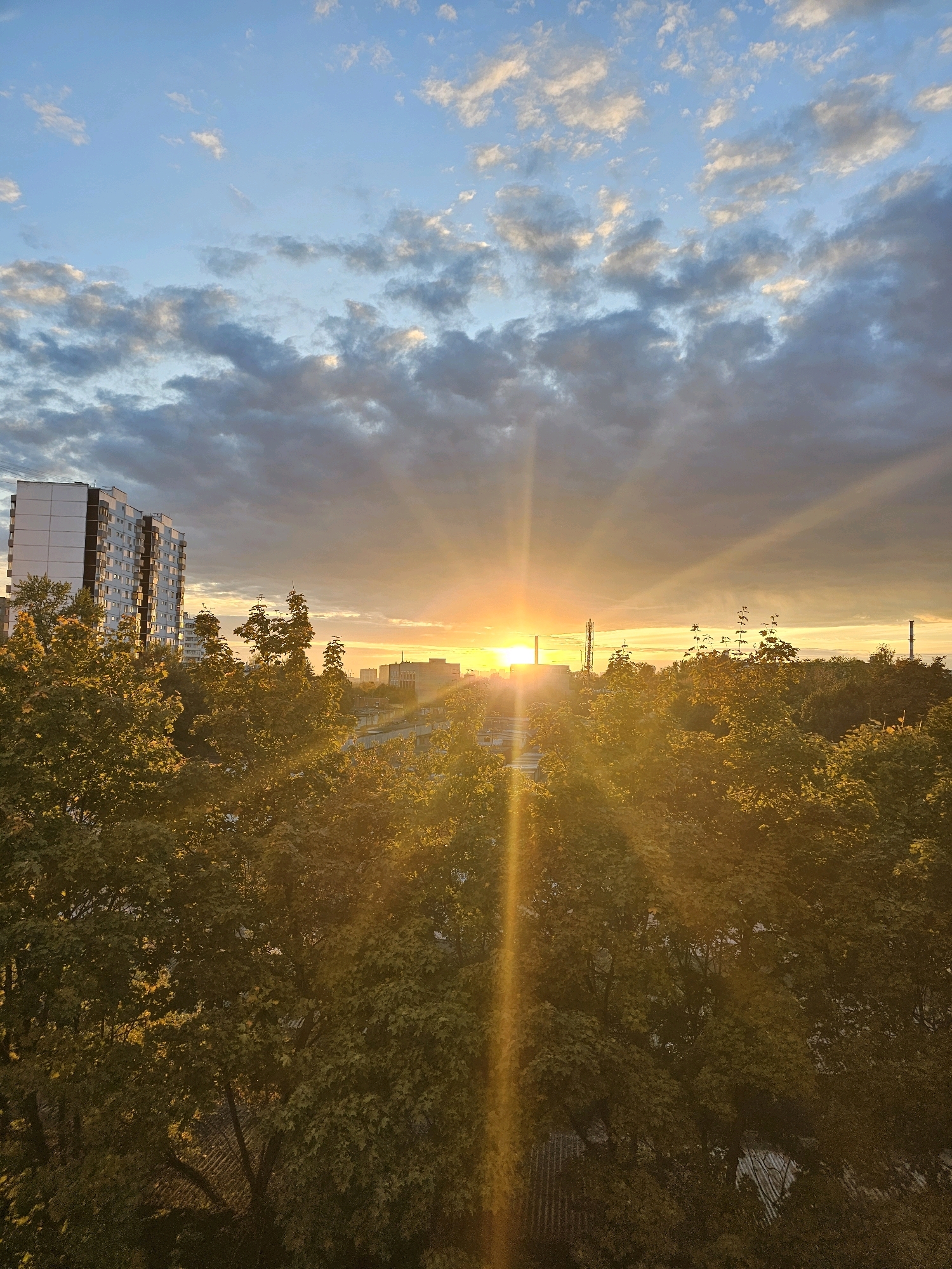 An aerial perspective captures a city skyline at sunset, featuring the distinctive architecture of Khimki, Russia. The sky, a canvas of blues and grays, is illuminated by the warm, golden rays of the setting sun, which burst through scattered clouds and create a dramatic sunbeam effect. In the foreground, lush green trees with autumnal hues frame the scene, their leaves catching the golden light. Beyond the trees, a tall, multi-story residential building, characteristic of Soviet-era architecture, stands prominently on the left. In the middle ground and distance, other buildings and industrial structures, including chimneys, dot the landscape, all bathed in the soft glow of the sunset. The overall atmosphere is one of serene twilight in an urban setting.