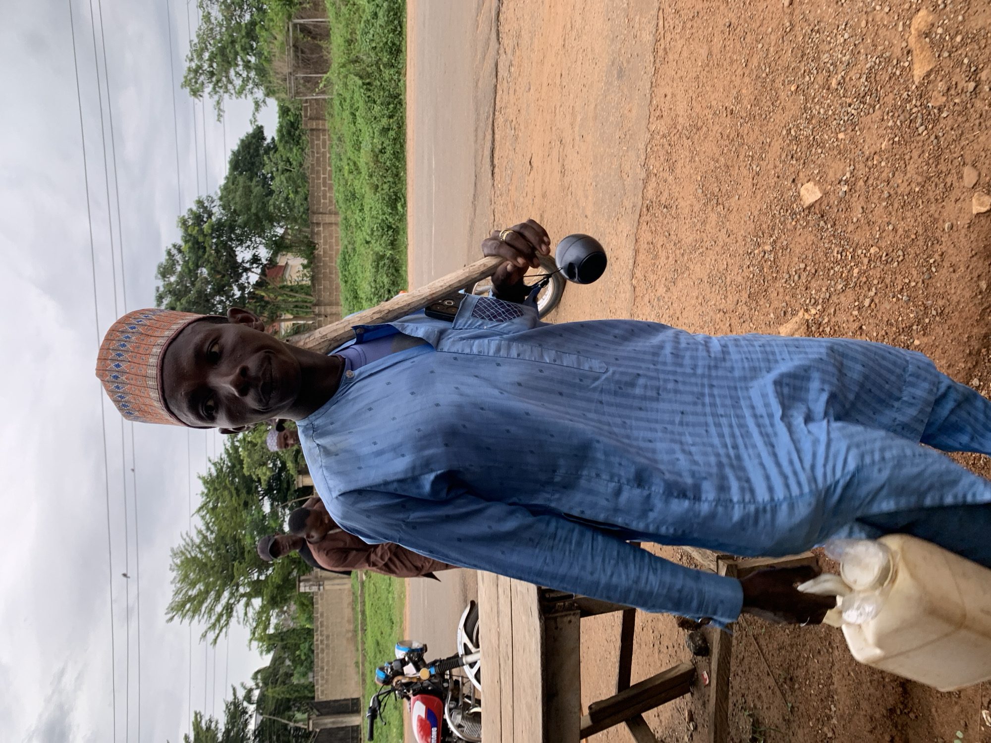 In Gbeleko, Nigeria, an outdoor scene unfolds under an overcast sky. The foreground features a dark-skinned man, approximately 30-40 years old, facing slightly right and looking towards the viewer's left. He wears a light-blue long-sleeved tunic and matching trousers, both patterned with subtle vertical stripes and small squares. A multi-colored cap, predominantly orange and blue, adorns his head. He carries a pale, off-white plastic jerrycan with two spouts in his left hand. A wooden staff rests on his right shoulder, from which a small, black, spherical object hangs by a cord. A dark-colored smartphone is visible in the pocket on his right side, beneath the staff. He wears a gold-colored ring on the ring finger of his right hand.
Behind the main subject, a group of men is visible. One man, positioned slightly left and rear of the main subject, wears a brown shirt and looks downwards. Further back, another man with a dark cap is partially obscured. To the far right, a third man also appears to be wearing a cap.
The ground consists of reddish-brown dirt and gravel. To the left, a rustic wooden table stands beside a red and white motorcycle with a blue headlight, parked near a paved road. Behind the individuals, a low concrete block wall is partially obscured by dense green vegetation. Beyond the wall and vegetation, a road is visible, suggesting a semi-urban or rural setting. The upper portion of the image shows an overcast, cloudy sky with overhead utility wires. Trees and lush greenery are scattered throughout the background.