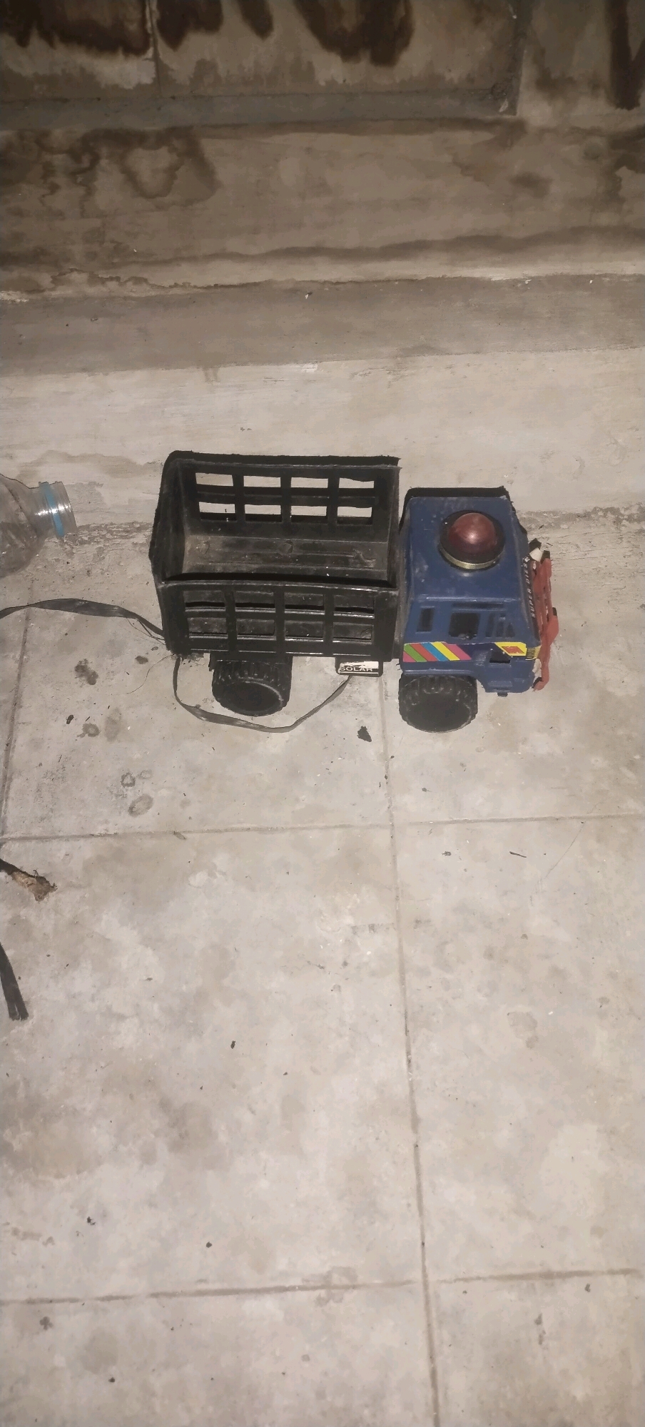 A toy truck, predominantly blue and black with some red accents, is positioned on a concrete floor. The truck appears to be a flatbed with a removable cargo section. A red light is visible on top of the cabin. There is a plastic bottle to the left of the truck, and a black strap extends from the truck towards the left side of the image. The floor is tiled with visible grout lines and has some stains and debris. The background consists of a textured concrete wall with dark water stains or discoloration. The scene appears to be indoors, possibly in a neglected or unfinished space, with no clear indication of the specific location in Daleman, Indonesia, beyond the provided context. There are no people or animals in the image.