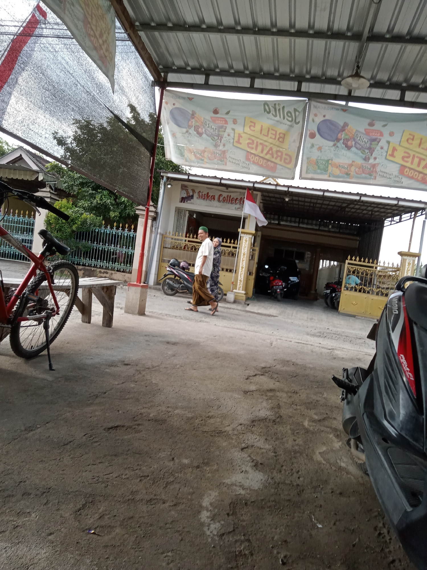 In the city of Simo, Indonesia, a scene unfolds under a corrugated metal awning. Two individuals, one man in a white shirt and brown pants and the other in a patterned dress, walk across a paved area. A red bicycle stands to the left, leaning on its kickstand. In the background, a building with a white facade and large open doorways reveals several motorcycles and a dark-colored car parked inside. Two large banners advertising "Jelly" are displayed prominently above the building. To the right, a yellow metal gate with ornate designs stands open, leading to another area with more motorcycles. A portion of a dark-colored scooter is visible in the foreground on the right side of the image. The ground is a rough, uneven asphalt surface. The overall atmosphere is that of an outdoor market or roadside establishment.