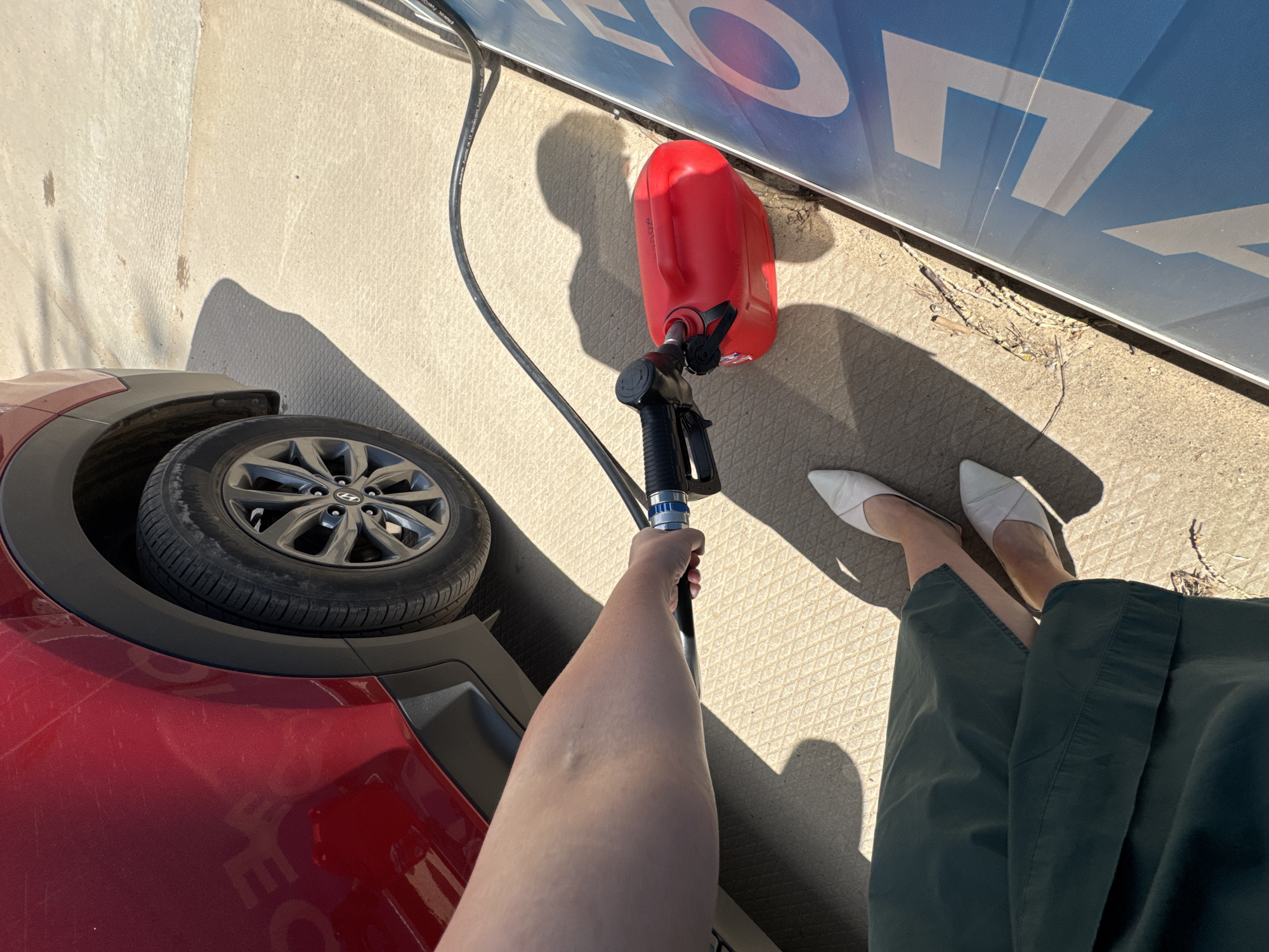 In Chaikovsky, Russia, a person, dressed in dark green wide-leg pants and white pointed-toe mules, stands on a textured concrete surface. The individual is holding a red portable fuel can with a black nozzle inserted into its opening. A black hose extends from the nozzle. The fuel can is positioned next to a large blue sign with white lettering, parts of which are visible. To the left, a portion of a red car is visible, including its rear wheel. The scene is outdoors, under sunlight, casting distinct shadows.