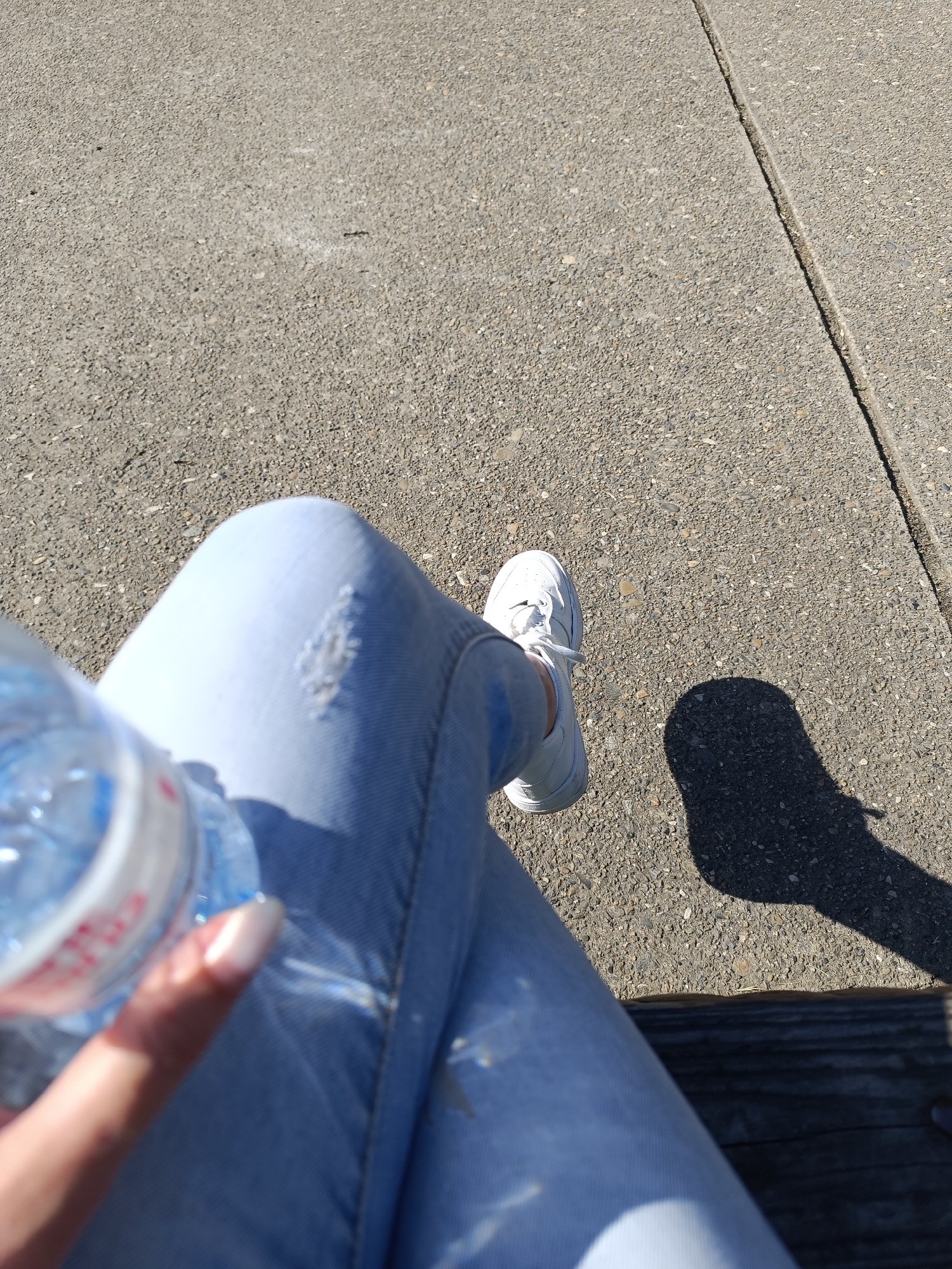 Here is a detailed description of the image:
The image appears to be taken from a first-person perspective, looking down at the legs of someone seated outdoors.
**People:** Only parts of a person are visible. The lower legs and feet are visible. A hand is holding a water bottle.
**Subjects:** The primary subjects are the person's legs, the water bottle, and the surrounding environment.
**Objects:** Key objects include:
* Light blue jeans.
* A single white sneaker.
* A clear plastic water bottle partially filled with water.
* Wooden bench.
**Actions:** The action is the person is sitting, holding the water bottle.
**Interactions:** The person is interacting with the bottle. The overall scene shows an individual taking a break outdoors.
**Environment:**
* **Surface:** A gray concrete or asphalt surface makes up the background. A narrow seam runs down the right of the frame.
* **Lighting:** The lighting appears to be bright and sunny, creating shadows.
* **Location:** The location appears to be a public space in Unknown, Unknown, given the concrete surface and the presence of a bench.