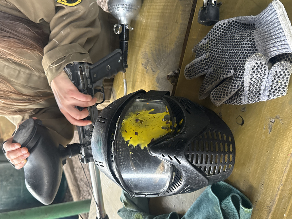 A paintball gun with a full hopper is held by someone wearing a tan jacket, positioned beside a paintball mask splattered with yellow paint. Gray gloves with black dots are resting on a tan surface next to the mask. A green cloth is visible at the bottom of the mask. The equipment suggests a paintball game or activity.