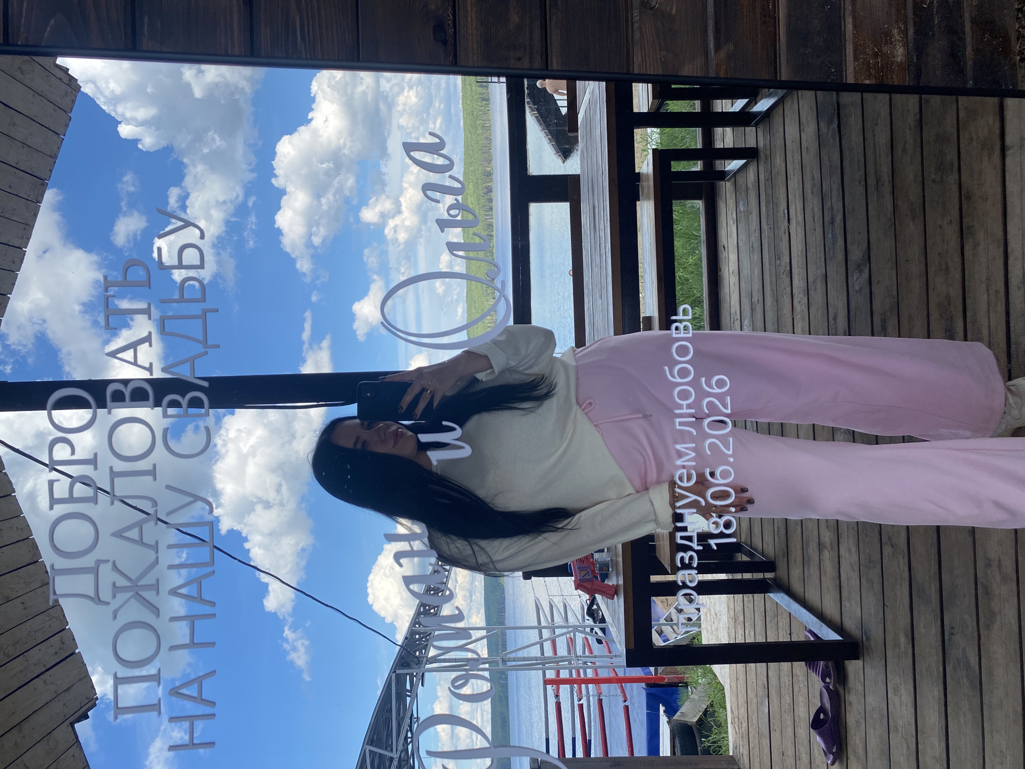 The media file is a portrait-oriented photograph taken in Chaikovsky, Russia. The primary subject is a young woman with long dark hair, dressed in a cream-colored sweatshirt and pink sweatpants. She is holding a smartphone, seemingly taking a selfie in a reflective surface, likely a mirror or a glass window. The reflection shows a clear blue sky with fluffy white clouds, and a body of water with trees lining the distant shore.
Text is prominently displayed across the reflection. On the left side, written vertically in large, white, serif font, are the words "ДОБРО ПОЖАЛОВАТЬ НА НАШУ СВАДЬБУ" which translates to "WELCOME TO OUR WEDDING". To the right of this, a name is written in a flowing, cursive font: "Ольга" (Olga). Further to the right, another line of text reads "Поздравляем с любовью 18.06.2026", translating to "Congratulations with love 18.06.2026".
The scene appears to be outdoors, possibly at a venue near a river or lake, given the water and tree line. The wooden planks visible in the foreground and background suggest a rustic or natural setting. The bright sky and clear clouds indicate fair weather, likely during the daytime. The overall mood conveyed is celebratory, with the wedding message and a sense of personal reflection captured by the selfie. The presence of a mobile phone and casual attire suggest a relaxed and informal event.