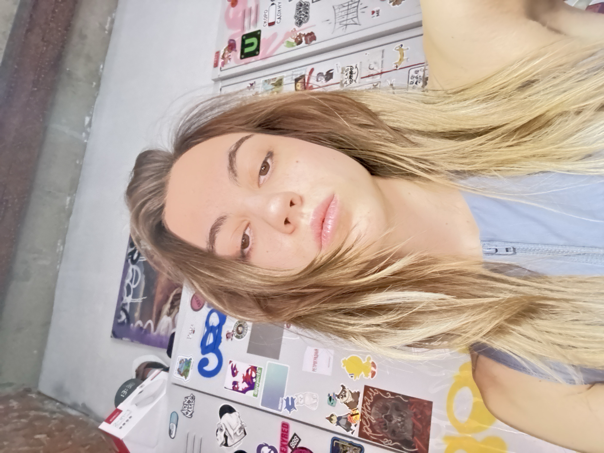 A young woman with blonde hair takes a selfie in front of a refrigerator covered in stickers. The refrigerator is white and appears to be in a kitchen or common area. The stickers on the refrigerator are varied, featuring cartoon characters, text, and abstract designs. The woman is wearing a light blue, zipped top. The background shows a textured wall and ceiling. There are no other people visible in the image.