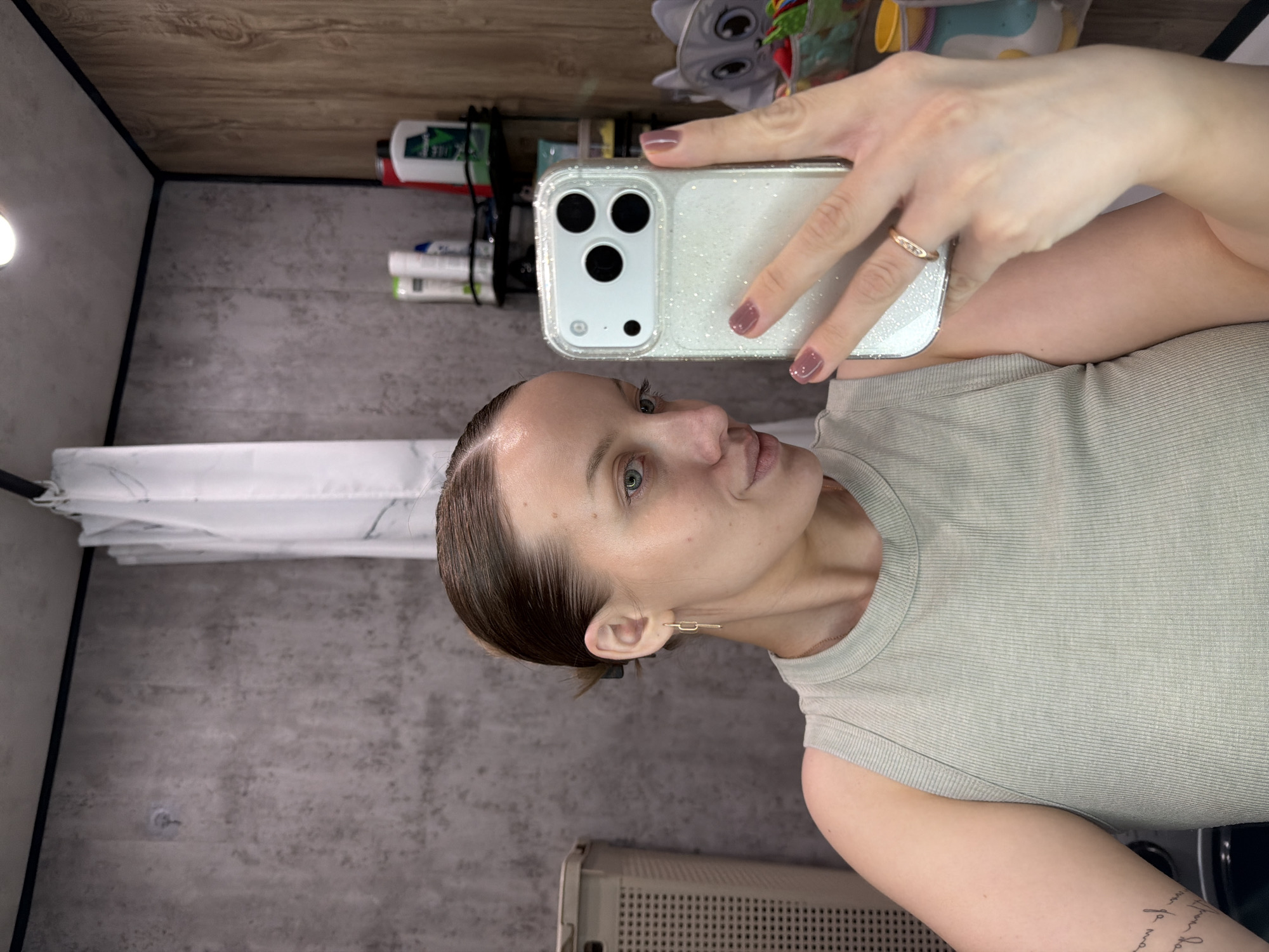 A medium shot selfie captures a woman holding an iPhone with a glittery clear case, featuring a triple camera setup, in her right hand. She is looking towards the camera with a subtle smile. Her dark brown hair is pulled back, revealing her ears with minimalist gold rectangular loop earrings. She has light blue or green eyes and visible freckles across her nose and cheeks. A gold ring with small stones is on her right ring finger, and her nails are painted a maroon-brown color. She wears a light olive or sage green ribbed tank top, and a script tattoo is visible on her left bicep.
The setting appears to be an indoor space, possibly a compact room or a camper/RV. The ceiling is paneled with light brown wood, while the walls have a mottled grey, textured finish. In the background, a shelf or ledge holds various personal care items, including bottles of Tresemmé and Herbal Essences products, a black comb, and a red and white electronic device. A bright light source shines from the upper left corner of the frame. To the lower right, part of a light-colored, possibly plastic, woven storage unit or appliance is visible. A white curtain hangs along the left wall. The location context for this image is Reshetnikovo, Russia.