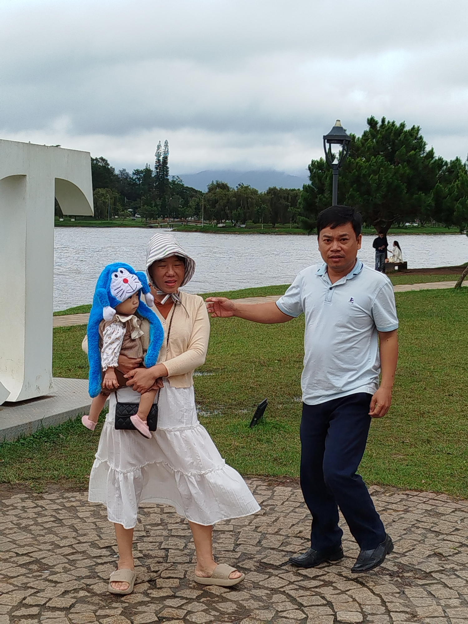 The image captures a family posing for a photo by Xuan Huong Lake in Da Lat, Vietnam. A woman on the left holds a baby wearing a blue cartoon-themed hat. She is dressed in a white dress and a light-colored cardigan. To her right stands a man in a light blue polo shirt and dark pants. They are on a cobblestone path with a body of water and lush green trees in the background, under a cloudy sky. A lamppost is visible near the man.