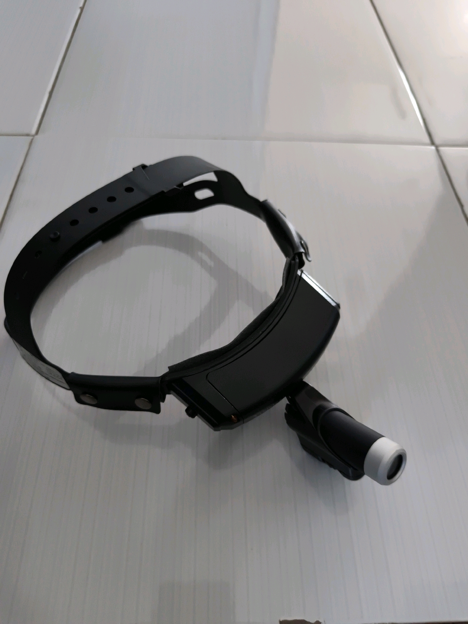 A black headlamp with a flashlight attachment rests on a tiled surface. The headlamp has an adjustable strap with several holes for size adjustment. The flashlight is cylindrical with a silver tip and is attached to the front of the headlamp. The tiles are white and arranged in a grid pattern. The lighting suggests an indoor environment with overhead light. No people are present in the image. The location is Tombatu Dua, Indonesia.