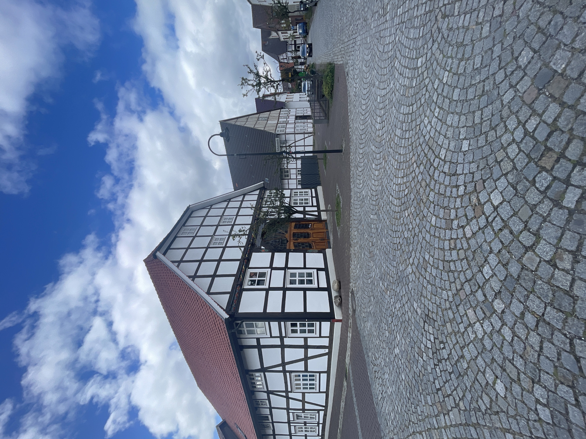 The image depicts a street scene in Salzkotten, Germany, featuring traditional half-timbered buildings. In the foreground, a cobblestone street curves towards the right. The prominent building on the left is a large, two-story half-timbered structure with a red tiled roof. It has white infill panels and dark timber framing, with numerous white-framed windows. Two rounded stones are placed near the corner of this building.

Further down the street, more half-timbered houses line both sides. A street lamp stands on the left side, with a street sign that appears to read "Schützenstr." A few cars are visible on the street, with one dark sedan in motion. The sky is partly cloudy, with a mix of blue sky and white clouds. The environment suggests a calm, perhaps historic, urban setting.