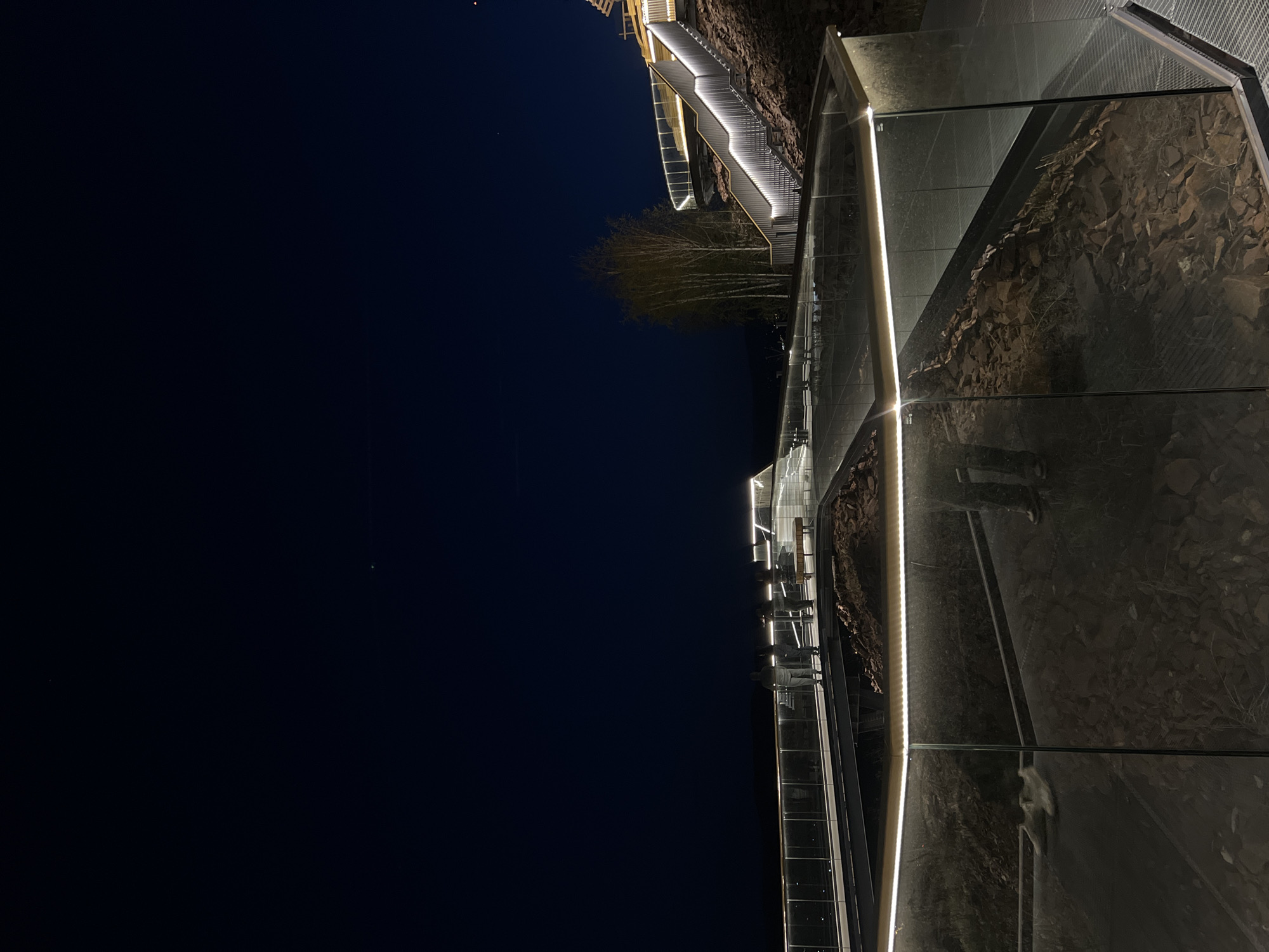 The image depicts a nighttime view of the "Torgashinsky Ridge" observation deck in Krasnoyarsk, Russia. A brightly lit staircase ascends a dark, rocky hillside. A glass-walled walkway runs along the edge of the hillside, reflecting the sky and the surrounding terrain. The sky is dark blue to black, with a few visible stars. The illumination highlights the modern design of the walkway and the rugged texture of the natural landscape.