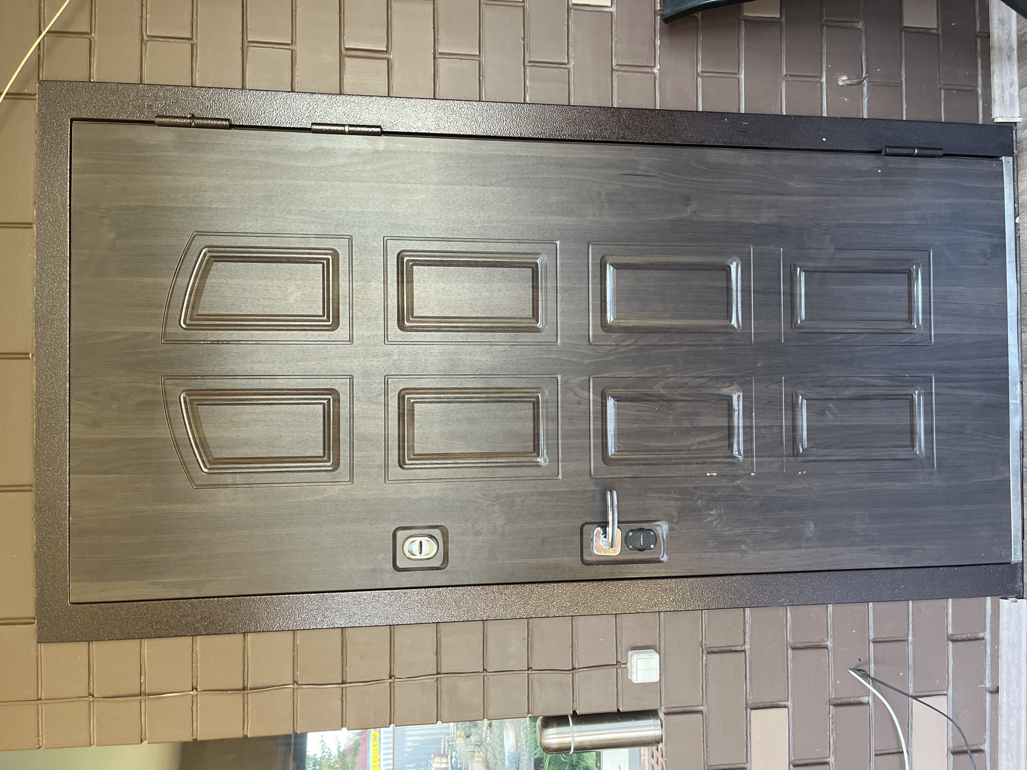 The image shows a dark brown, wooden-grained metal door with a textured dark brown metal frame. The door has six recessed panels, two on the top row with arched tops and four rectangular panels below. A silver door handle and a silver lock mechanism are visible on the right side of the door. A circular peephole or lock cylinder is present above the handle. The door is set into a wall constructed of light brown bricks. To the left of the door, on the brick wall, is a silver thermal flask and a white electrical outlet. A thin white electrical wire runs from the outlet up the wall and over the door frame. A window is partially visible to the far left. The lighting suggests it is daytime. The scene is an exterior view of a building entrance. The location is identified as being in Saratov, Russia.