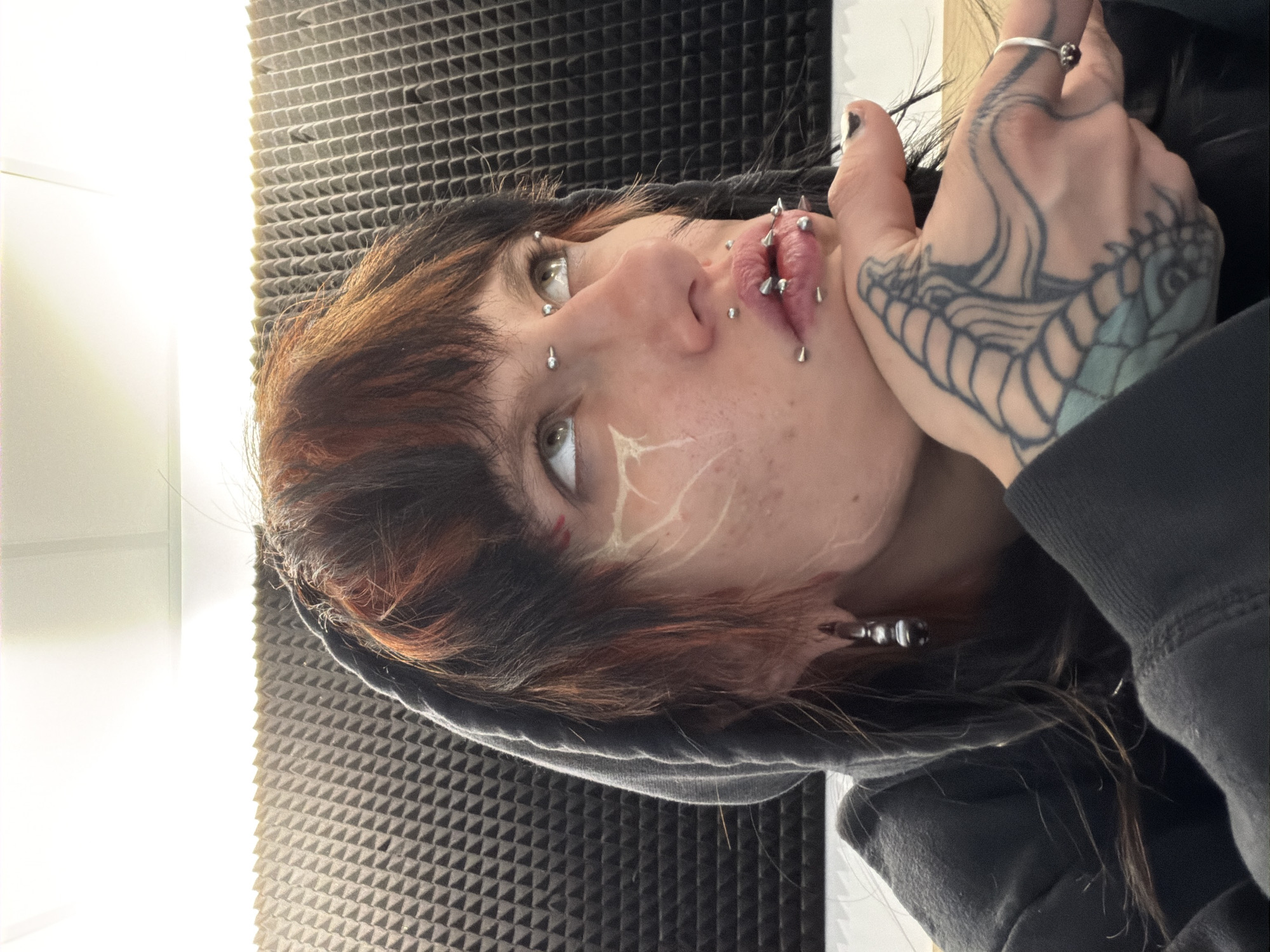 A person with multiple facial piercings, including lip and cheek piercings, and a tattoo on their right hand, is shown in an indoor setting. The person is wearing a dark hooded sweatshirt. Their hair is dark with red highlights. They have light skin with what appears to be either a scar or painted design on their cheek. The background includes acoustic foam paneling and a white wall. The city of Sofia, Bulgaria, is not discernible from the provided image.