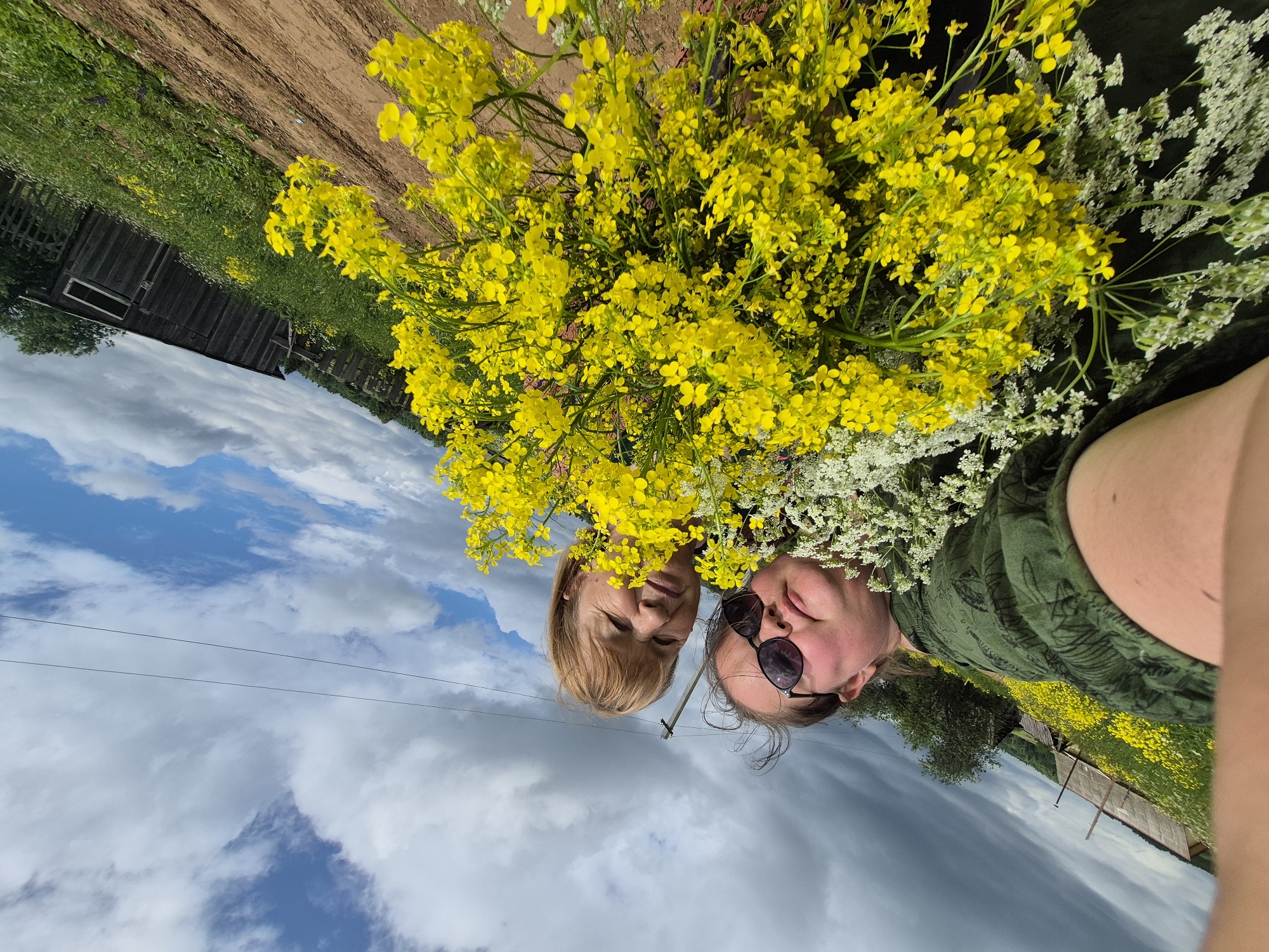 This is a selfie taken outdoors, tilted at an upward angle, featuring two people and a large bouquet of yellow and white flowers. The scene appears to be a rural or countryside location, possibly near fields of yellow flowers, with a wooden structure and fence visible in the background. The sky is partly cloudy with visible power lines. The person in the foreground is wearing sunglasses and a green patterned top. The person behind them has blonde hair and is looking towards the camera. The image conveys a casual, outdoor setting.