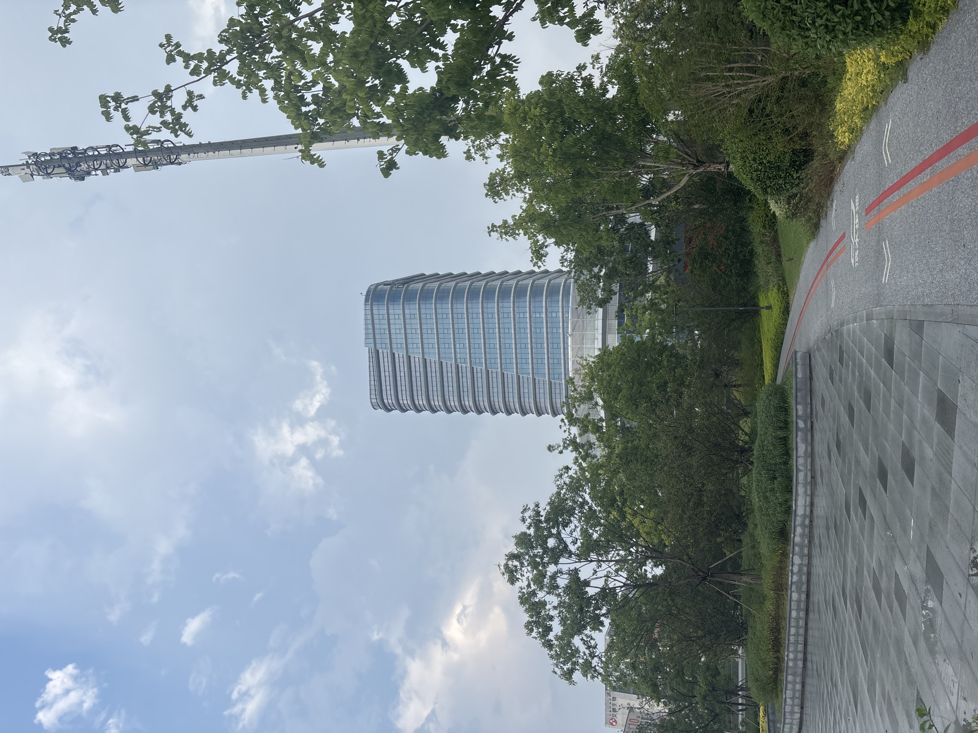 The image depicts a modern park area in Suzhou Industrial Park, China, featuring a paved walkway with red and orange lane markings, lush green trees, and a contemporary glass-facade building in the background. A tall telecommunications tower is visible on the right side of the frame. The sky is overcast with some clouds, suggesting a temperate day. The ground appears damp, possibly from recent rain. There are no people visible in the scene.