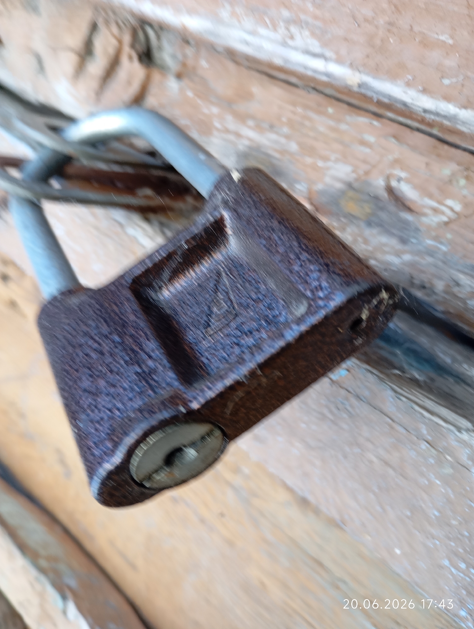 The image shows a close-up, high-angle view of a rusty padlock securing a wooden surface, possibly a door or gate. The padlock's shackle is visible and appears to be made of metal. The wooden surface exhibits signs of wear and tear, with visible grain and some peeling paint. The lighting suggests it is daytime, but the specific location within Урыцкі сельскі Савет, Belarus, cannot be determined from the image alone. There are no people or animals present in the scene. The date stamp "20.06.2026 17:43" is overlaid on the bottom right corner of the image.
