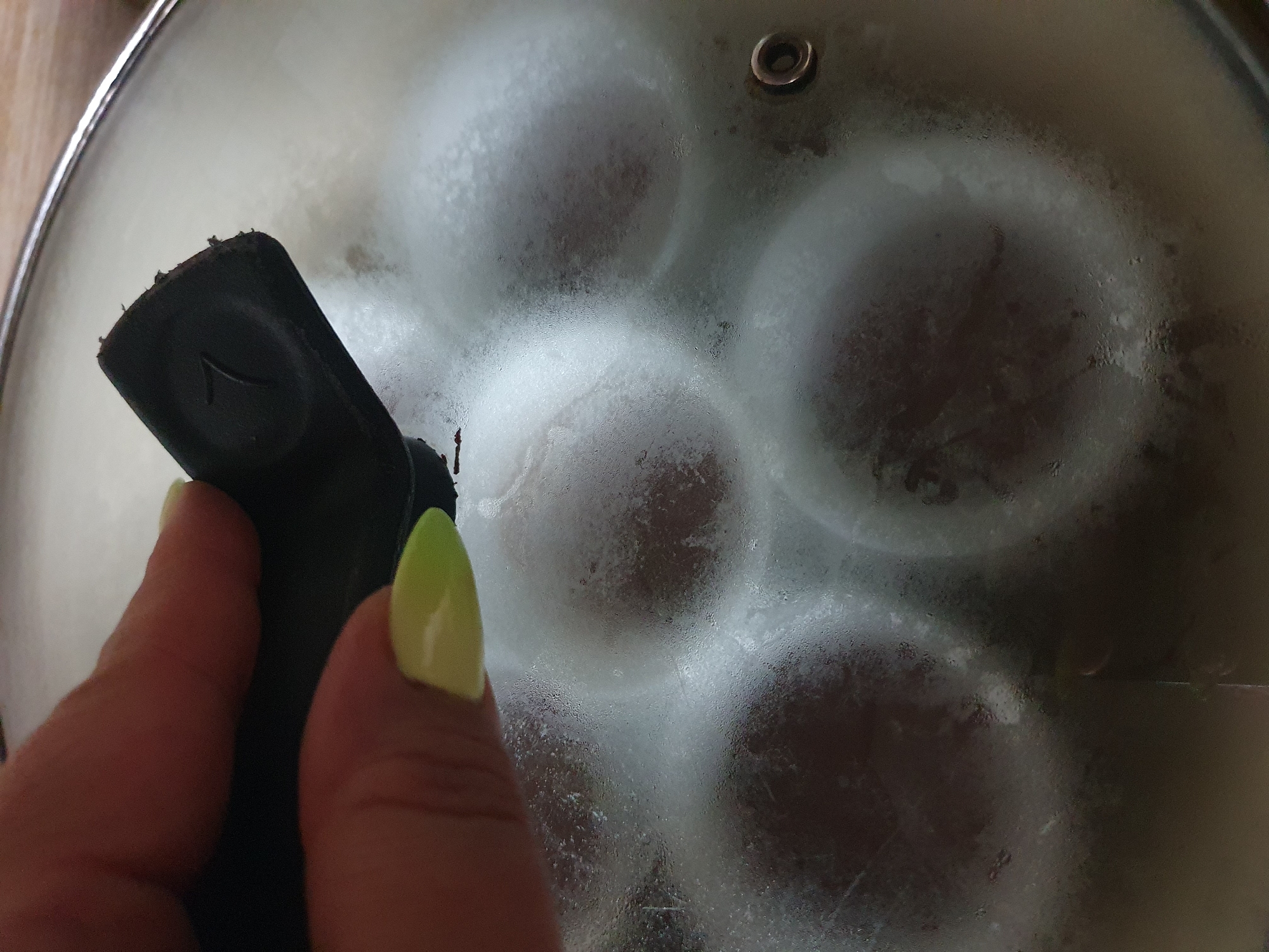 A close-up shot shows a hand with lime-green nails holding a black pot lid with a raised number "7" molded into its handle. The lid is placed over a pot, and condensation is visible on the underside of the glass, obscuring the contents within. The pot appears to contain several round, dark-colored items, possibly food. The background is indistinct and appears to be a kitchen environment. The image is taken in Shiraz, Iran.