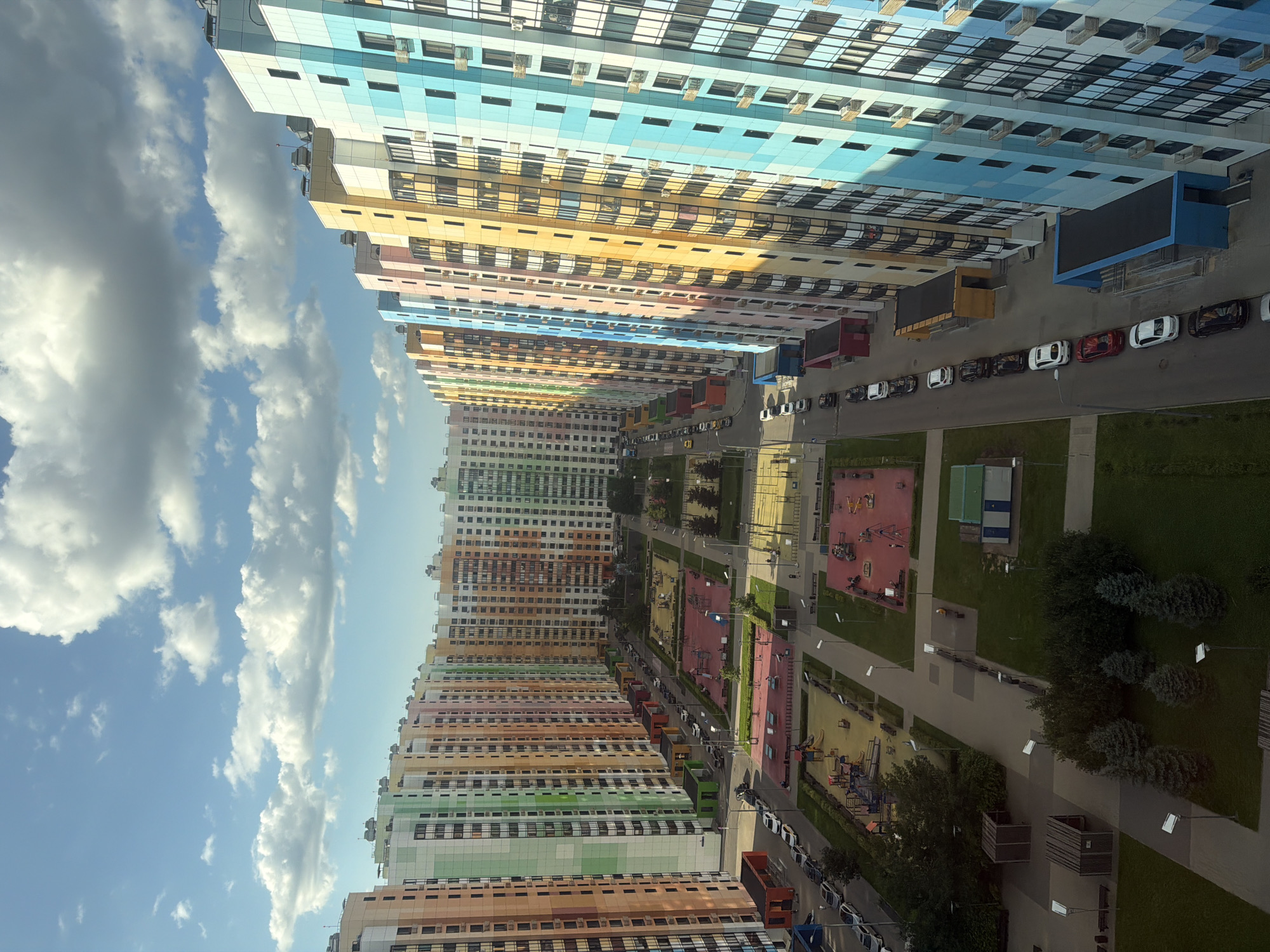 The image shows a view from above of a residential complex with brightly colored buildings and a clear sky with scattered clouds. The buildings are tall, multi-story structures with facades in a variety of pastel colors, including pink, orange, yellow, green, blue, and teal. They are arranged in a curved row, creating a sense of depth and perspective. Between the buildings, there are courtyards with playgrounds, green spaces, paved walkways, and parked cars. The playgrounds feature swings, slides, and climbing structures. Several cars, mostly sedans, are parked along the streets and in parking areas. A few people are visible walking or standing in the courtyards and on the sidewalks. The overall impression is one of a modern, well-maintained urban neighborhood with a vibrant and cheerful aesthetic.