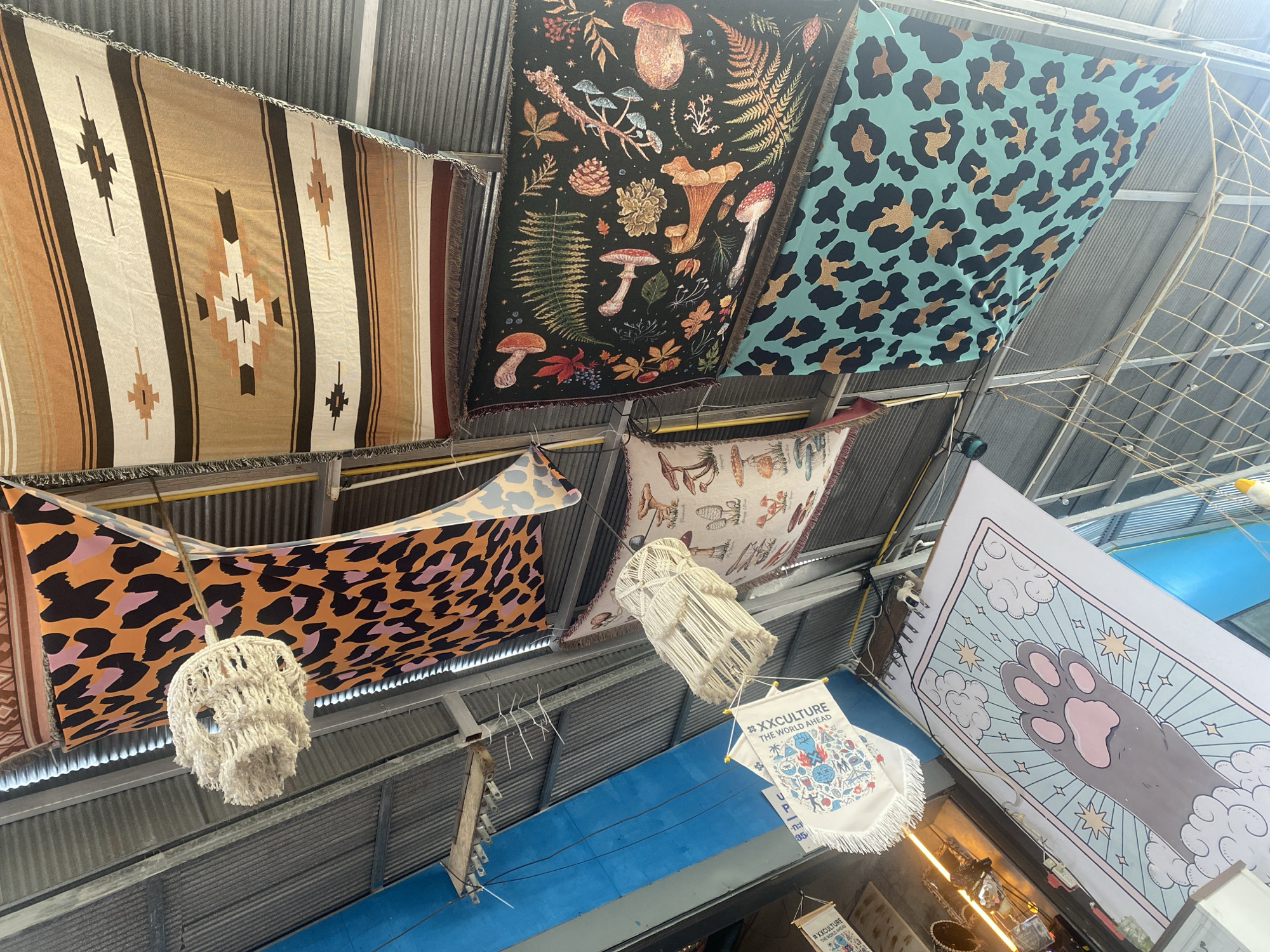 The image, taken from a low angle, shows the ceiling of a building, possibly a market or fair, in Baan Nai Suan, Thailand. The ceiling is made of corrugated metal. A series of tapestries or blankets are suspended horizontally across the ceiling, creating a decorative effect. The tapestries feature various patterns: a striped design in earthy tones, a dark tapestry with botanical illustrations including mushrooms, a teal tapestry with a leopard print, and an orange tapestry with a similar leopard print. Underneath the tapestries hang decorative items, including macrame-style light fixtures and banners displaying the text "#XXCULTURE THE WORLD AHEAD" along with illustrations. In the lower right corner, a large poster depicting a cartoon cat paw surrounded by clouds and stars is visible. The overall scene suggests a bohemian or artistic aesthetic.