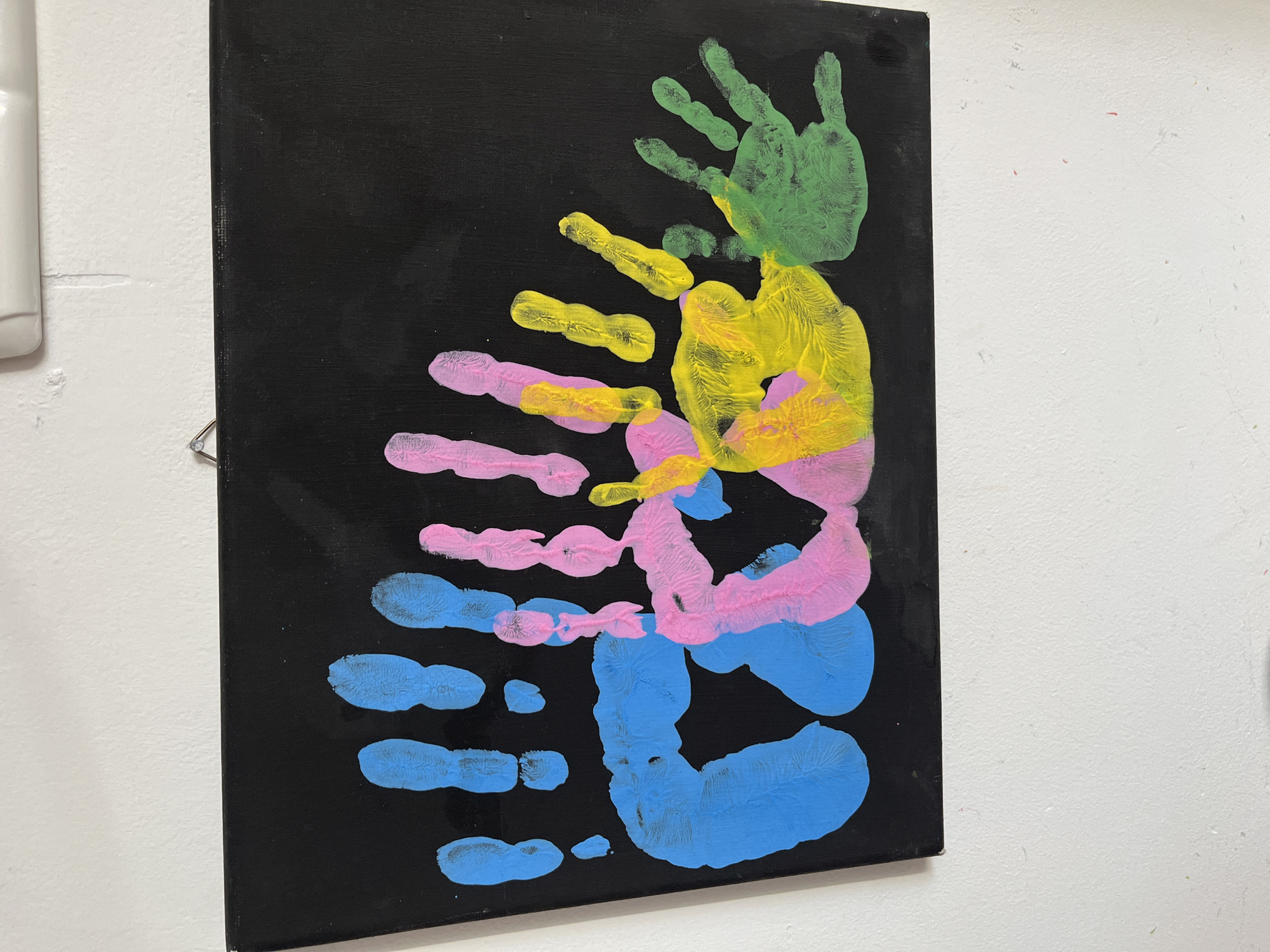 The image displays a black canvas with colorful handprints. The handprints are in various colors including blue, pink, yellow, and green. There are several handprints, some are fully formed, while others are partial. The handprints overlap and are arranged in a seemingly random pattern across the canvas. The canvas is hanging on a white wall, and a portion of a white ceiling fixture is visible at the top of the image. The scene appears to be indoors, likely in a residential or art studio setting. There is no information provided to indicate the location is Kohtla-Järve linn, Estonia.
