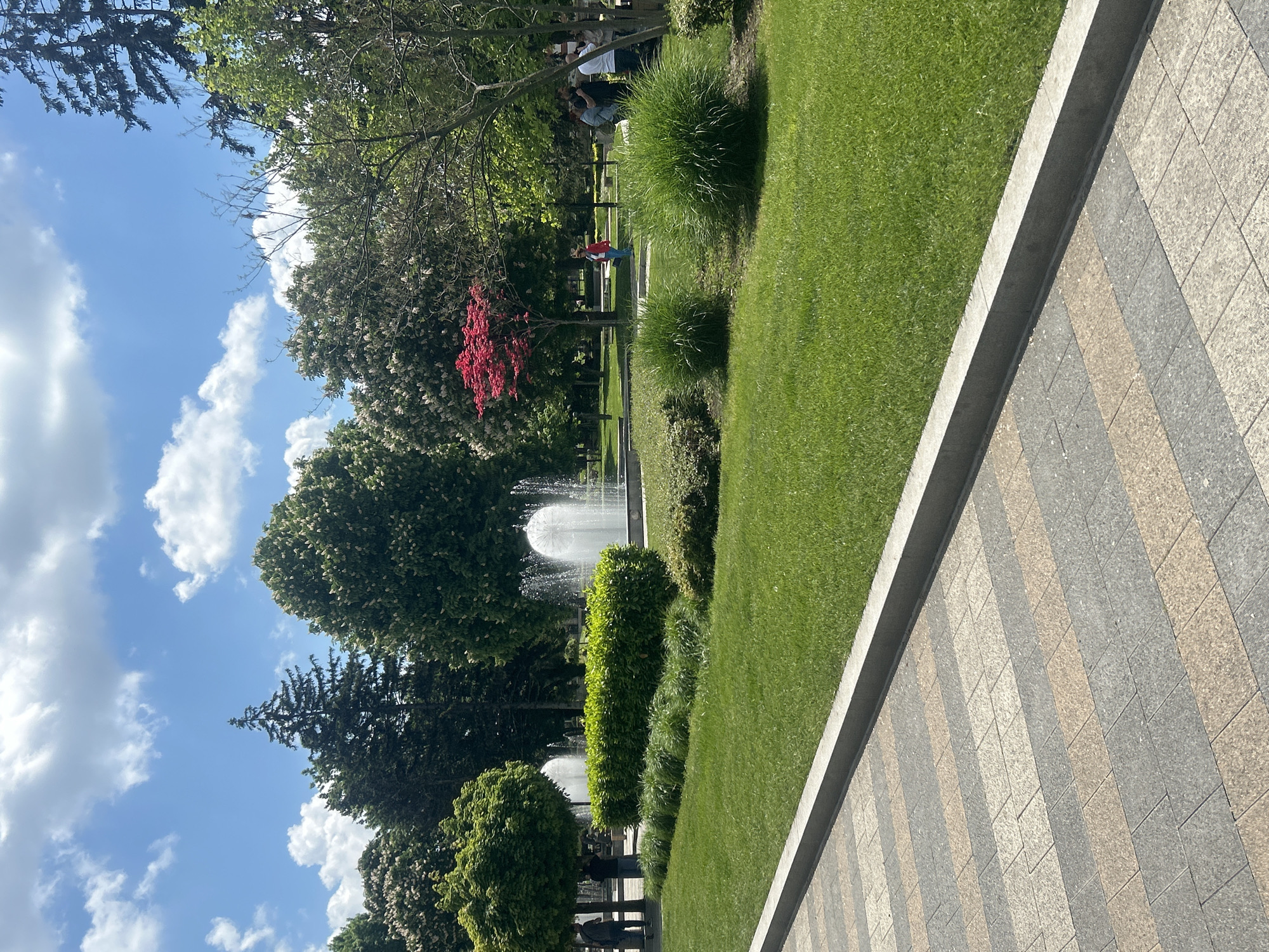 The image depicts a park scene in Pleven, Bulgaria, featuring a walkway composed of gray and beige rectangular pavers, bordered by a concrete edge. To the left of the walkway is a well-maintained lawn with various green plants, including shrubs and ornamental grasses. In the background, there are trees with dense foliage, including a tree with red leaves, partially obscuring two water fountains. A metal fence is visible behind the fountains. Several people are visible in the background, near the metal fence. The sky is visible in the upper left portion of the image with scattered white clouds.