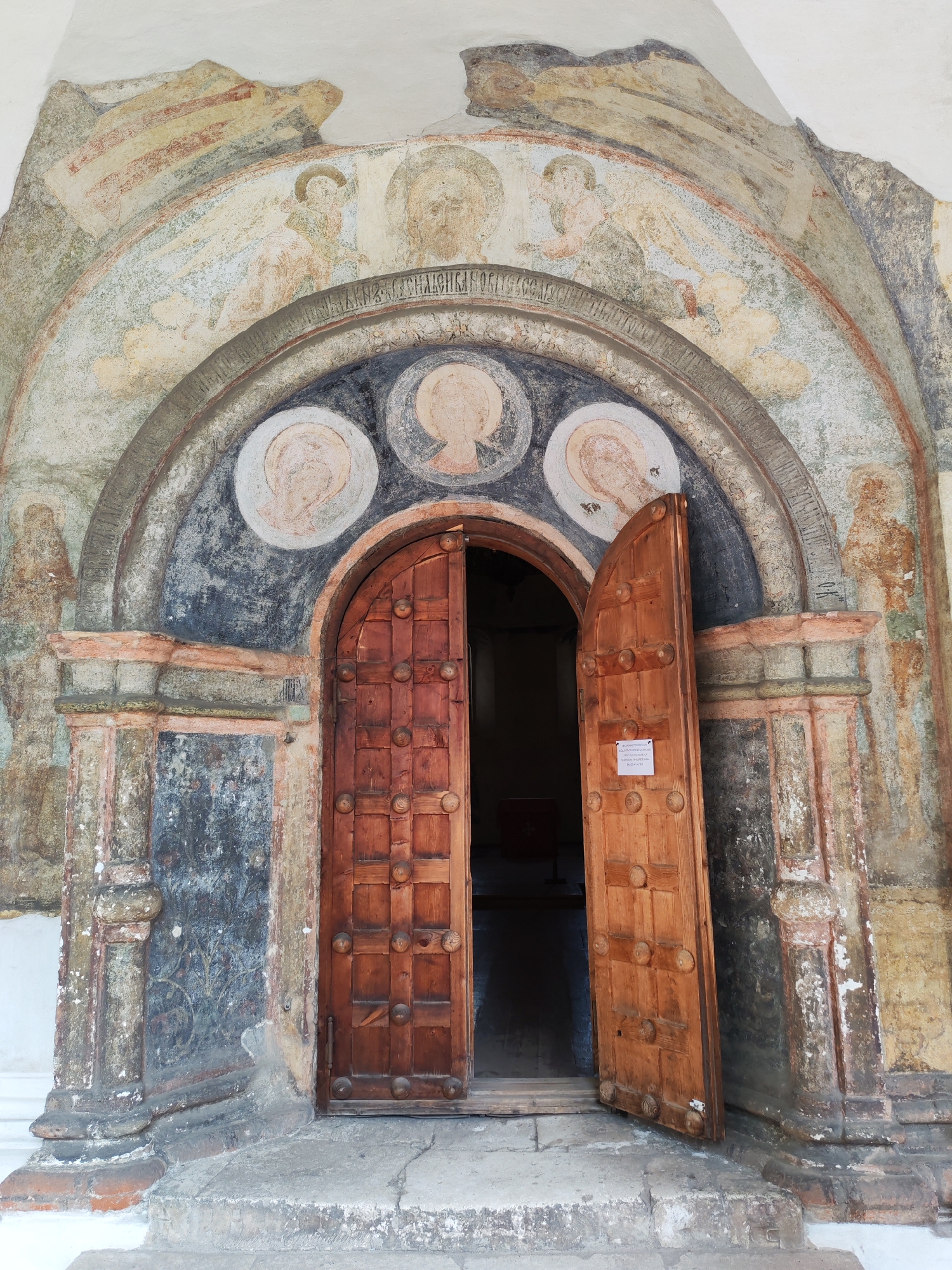 An open, weathered, double wooden door is centered in an arched entryway. The door is made of vertical planks with rows of decorative circular wooden bosses. The entryway is framed by stone and adorned with faded frescoes depicting figures and possibly religious scenes. The frescoes are multi-layered and show signs of significant age and wear, with colors like ochre, pale blue, and red visible. The architecture appears ancient, with crumbling plaster and weathered stone. The ground in front of the door is made of worn stone steps. A small white sign is attached to the inside of the right door leaf. The overall scene suggests a historical or religious site. This image was taken in Rusanovo, Russia.