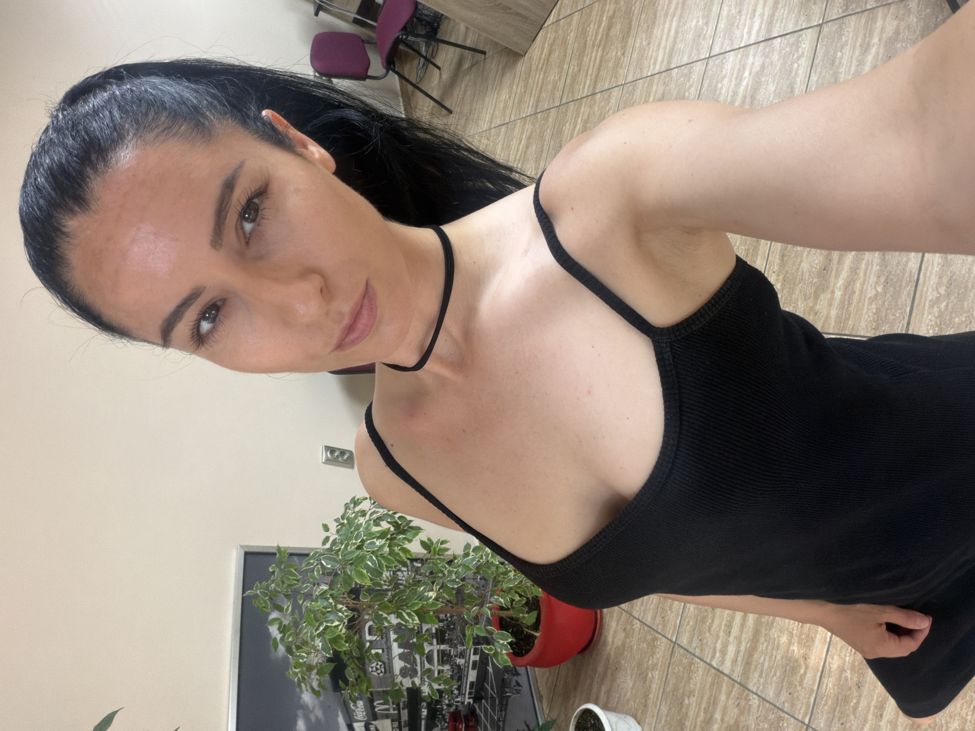 The image shows a woman in a black tank top and black choker necklace, posing for a selfie in an indoor environment. She has dark hair pulled back in a ponytail and is looking towards the camera with a slight smile. In the background, there is a potted plant with green and white leaves, a framed picture on the wall, and two purple chairs. The floor is tiled. The setting appears to be an office or a home. The image's metadata indicates it was taken in Pleven, Bulgaria.