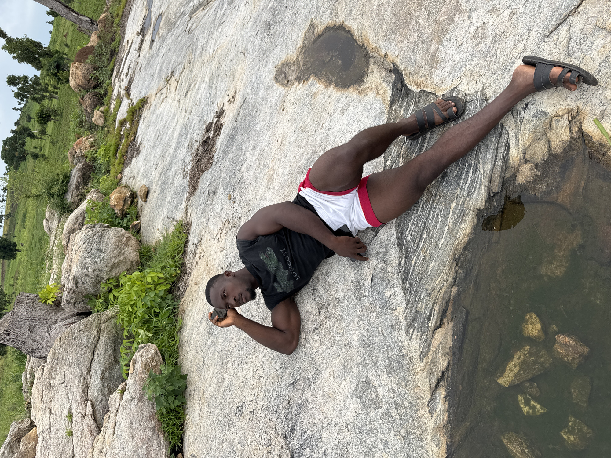 A man of African descent is lying on a large, weathered rock formation. He is wearing a black t-shirt with a green logo and white shorts with red trim. His head is resting on his left arm, and he is looking off to the right. The rock is covered in lichen and has cracks and crevices. In the background, there are more rocks, green vegetation, and a hill with scattered trees. The scene appears to be in a natural, outdoor setting. This location is identified as being in Akwanga, Nigeria.
