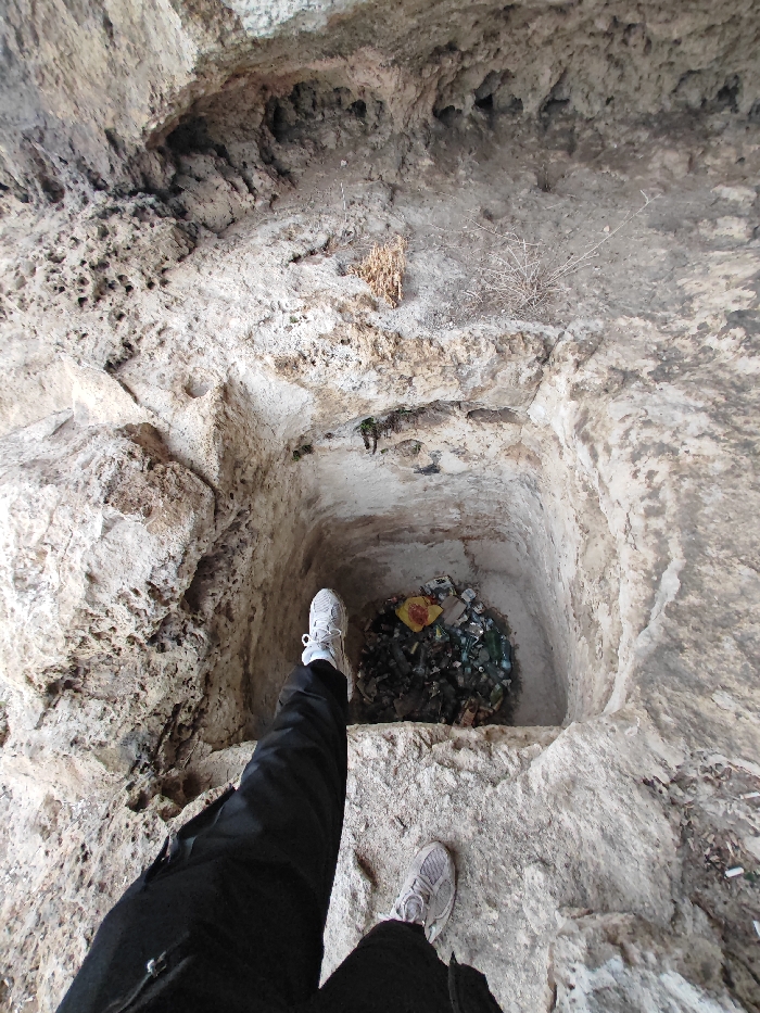 An overhead perspective photograph taken in the city of Shumen, Bulgaria, captures a deep, approximately square-shaped pit carved into light-colored, heavily textured rock. The pit is largely filled with refuse, including numerous clear, green, and brown glass bottles, plastic containers, and a distinctive yellow plastic bag or wrapper. In the foreground, the lower legs and feet of an individual are visible, wearing black trousers and white and grey athletic shoes, positioned at the edge of the pit, suggesting the person is looking into it. The surrounding rocky environment displays natural weathering, featuring various indentations, small holes, and ledges, with sparse dry vegetation present on some surfaces.