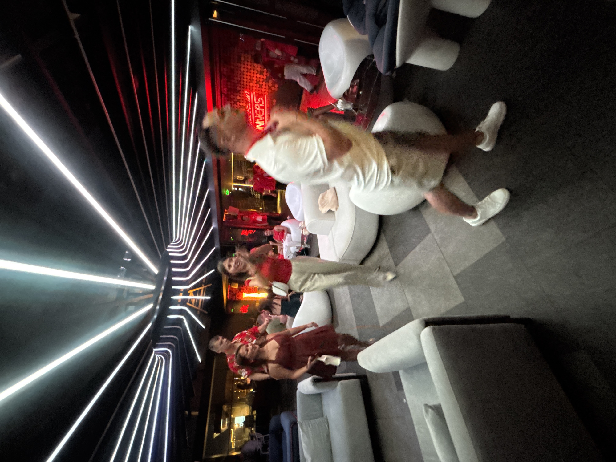 In a dimly lit, modern establishment with a futuristic ambiance, a group of young adults appears to be enjoying a night out. The scene is characterized by sleek, white, modular furniture arranged in seating areas, and a striking architectural feature of parallel, illuminated lines, possibly neon or LED strips, creating a tunnel-like effect.
The subjects are several individuals, some of whom are casually dressed and interacting. In the foreground, a woman in a dark red dress is holding a clutch bag, and a man in a red floral shirt is standing next to her. Further back, a woman in a red top and light-colored pants is animatedly gesturing, and another person in a white t-shirt and light shorts is seated on a white modular sofa, facing away from the camera. The overall atmosphere suggests a lively social gathering, possibly a party or a night at a bar or club.
The background reveals more of the establishment's interior, including a wall with red patterned lighting and visible text that appears to be part of a neon sign, possibly saying "WINNERS". The lighting is dynamic, with the white and red lights creating a vibrant and somewhat disorienting effect. The time of day is clearly nighttime, and the indoor setting suggests an urban location, consistent with Đà Nẵng, Vietnam. The people's postures and expressions suggest a mood of enjoyment and social interaction.
