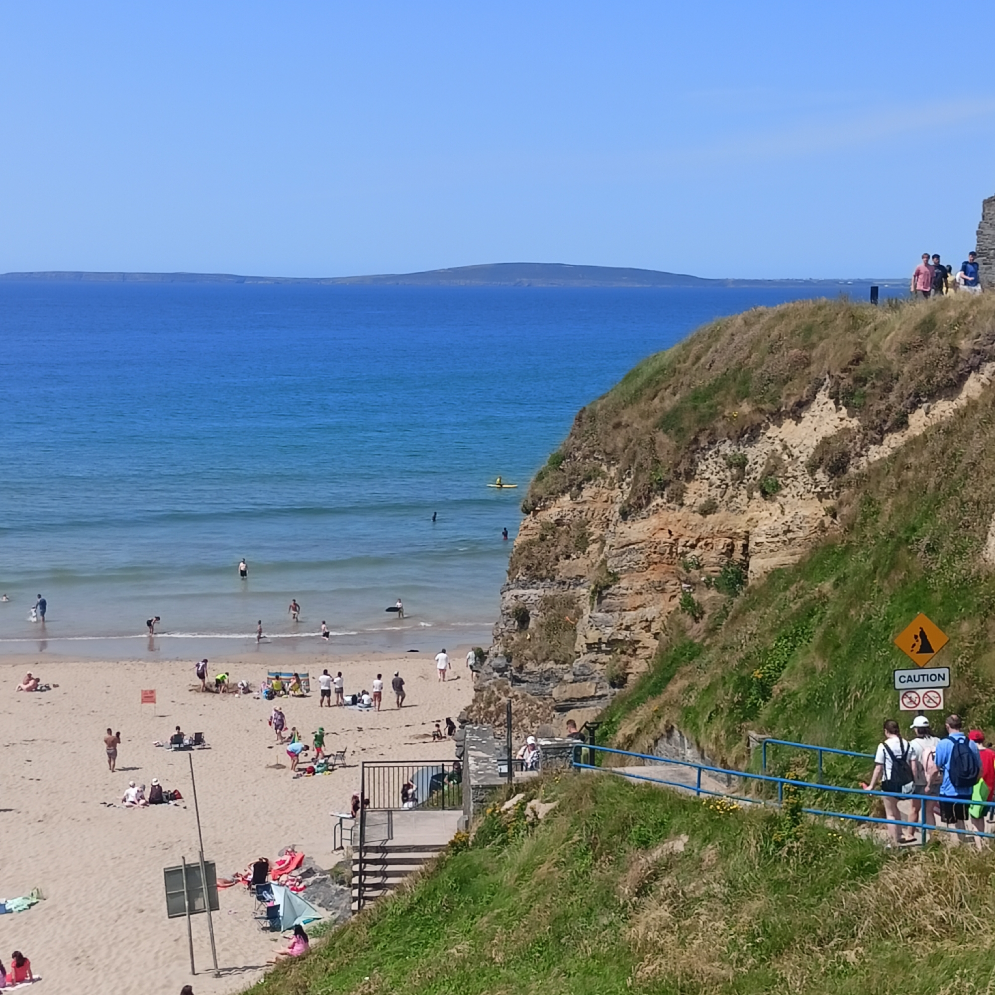The image captures a coastal scene at Ballybunnion, Ireland, on a clear, sunny day. In the foreground, a steep, grassy cliff face dominates the right side of the frame, with a paved pathway and a blue railing following its contour, indicating a scenic walking route. A "CAUTION" sign with a falling rocks symbol and a "no swimming" symbol is visible on the cliff face.
Below the cliff, a sandy beach stretches out, populated by numerous beachgoers. People are seen sunbathing on towels, building sandcastles, wading in the shallow waters of the ocean, and playing near the shoreline. The ocean itself is a vibrant blue, with gentle waves lapping at the shore. In the distance, a long, low-lying island or peninsula is visible on the horizon under a clear blue sky.
Several people are walking along the path on the cliff, descending towards the beach. One person is seen kayaking further out in the sea. The scene overall conveys a typical summer day at a popular seaside location.
