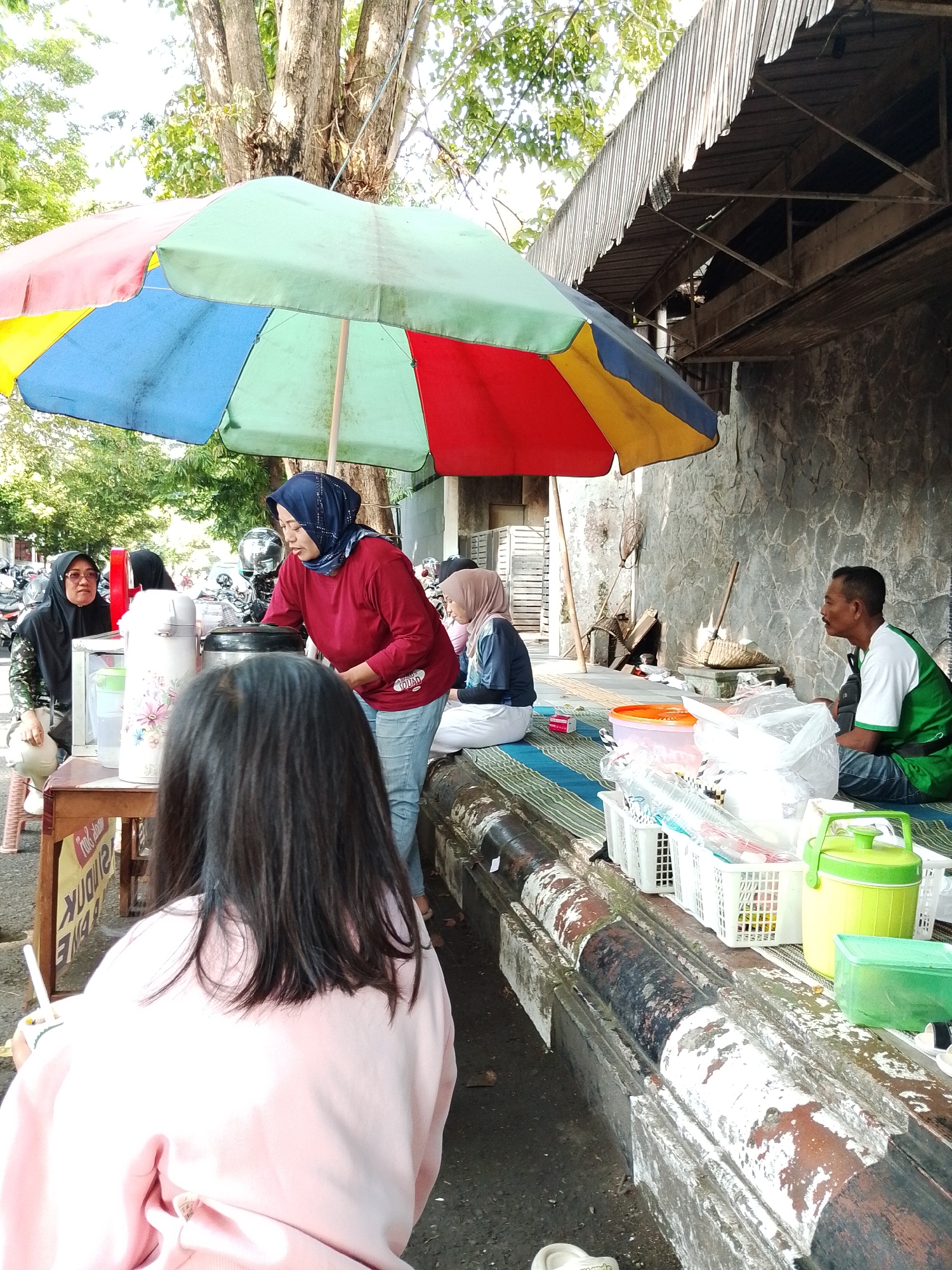 The image shows a street-side vendor in Kecamatan Pati, Indonesia. A woman wearing a maroon shirt and a blue hijab is serving a customer, whose back is to the camera, wearing a pink sweatshirt. The vendor is standing behind a small table with drink dispensers. A colorful umbrella provides shade. Two women wearing hijabs are seated nearby. A man wearing a green and white shirt is sitting on a mat. Various containers, baskets, and supplies are arranged along a ledge. The background shows a stone wall and a roof overhang.