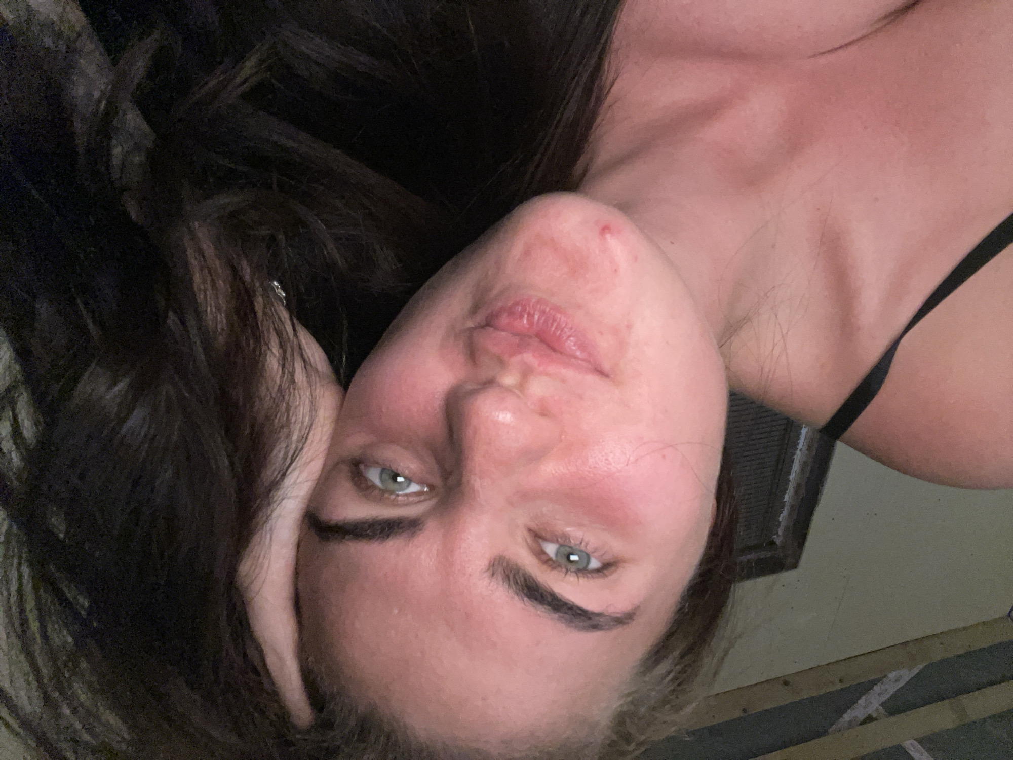 The image is a close-up, selfie-style photograph of a young woman with dark hair and light-colored eyes. She is lying down, with her head propped on her hand, and her hair is spread around her face and shoulders. The woman is wearing a black camisole or bra strap is visible. Her face is turned towards the camera, and she has a playful expression, with her lips pursed. There is a small red mark or pimple on her chin. The background appears to be indoors, with some wooden beams and a wall visible, suggesting a rustic or unfinished interior space. The lighting is dim, creating a warm and intimate atmosphere. The image does not contain any specific visual cues that can be used to determine its location in the city of Мосметаллоконструкция Онт, Russia.