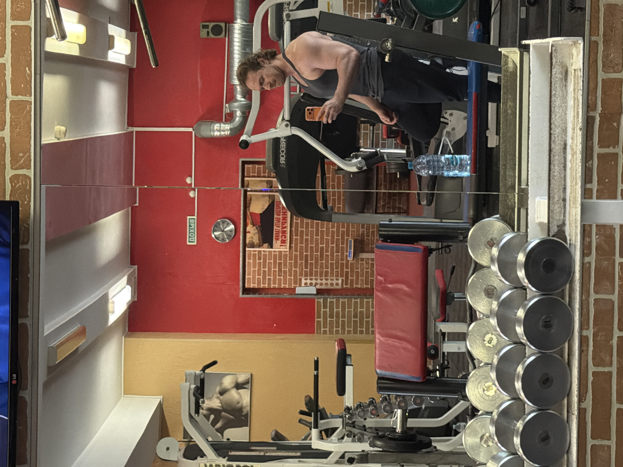 The image depicts the interior of a gym, likely in Saint Petersburg, Russia, judging by the Cyrillic script. A man wearing a tank top and dark pants is using a treadmill, looking at his phone. A bottle of water is visible on the treadmill console. Several weightlifting machines are present, along with a rack of dumbbells. The gym's walls are painted red and tan, with brick accents. A poster featuring a bodybuilder is on the wall. The scene is reflected in a mirror, causing a distorted perspective.