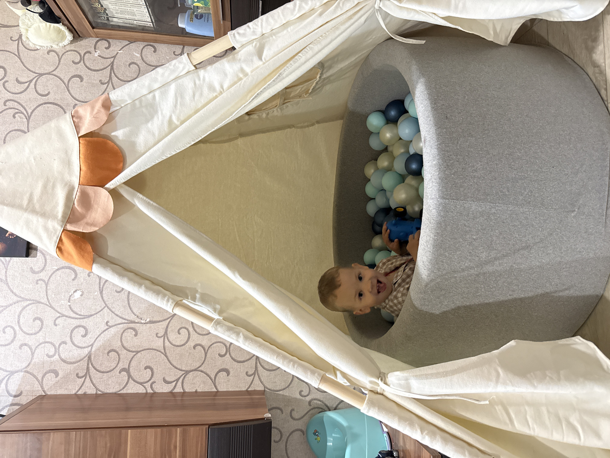 A low-angle, interior shot shows a baby boy in a gray ball pit, which is contained within a teepee-style play tent. The ball pit is filled with blue, gray, and white plastic balls. The boy is holding a blue toy. The teepee has white canvas walls with colorful fabric flower accents. The floor and surrounding furniture suggest an interior setting. The image was likely taken in Rusanovo, Russia.