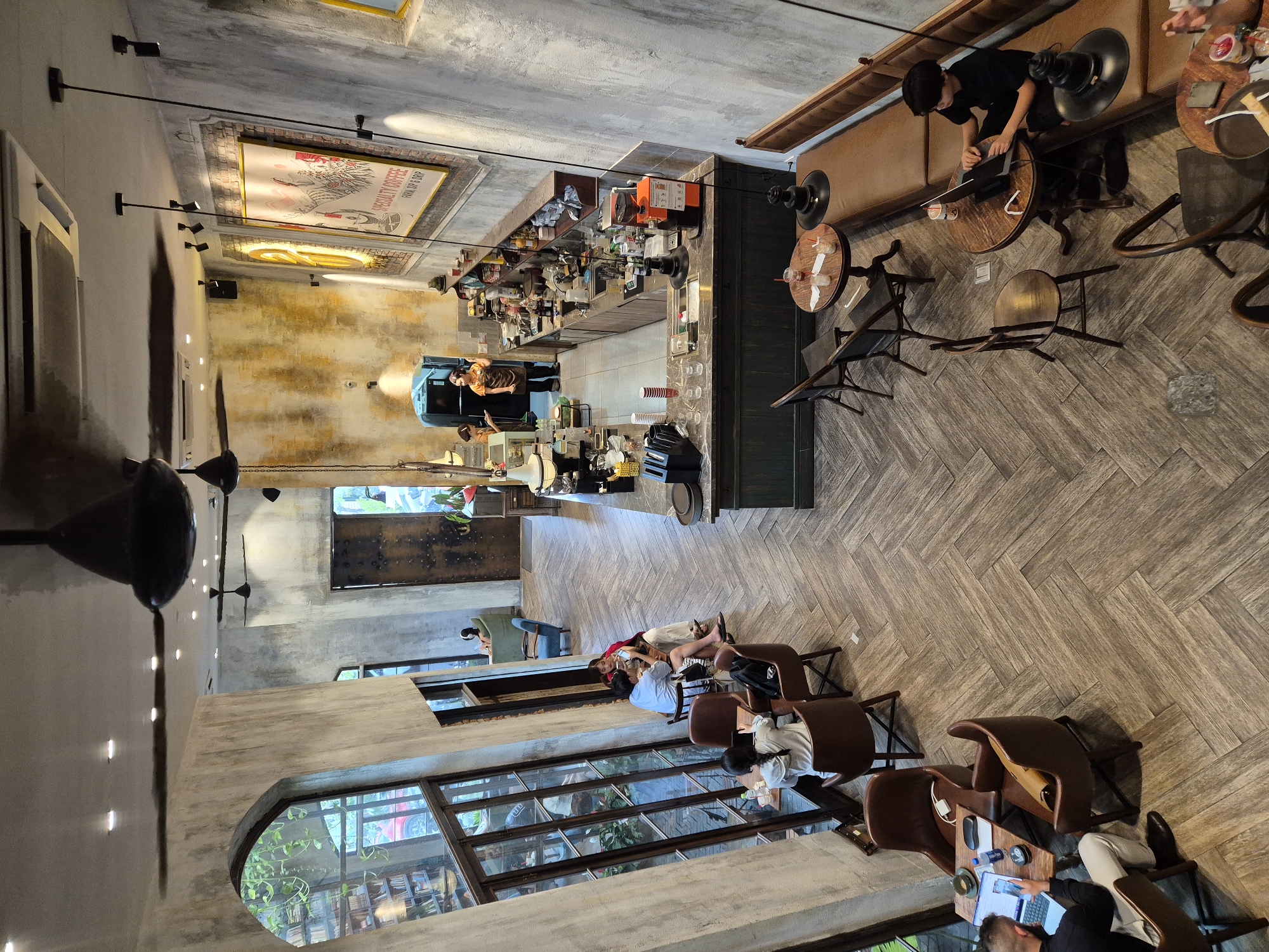 The image captures an indoor scene, likely a modern cafe with a rustic and industrial aesthetic, located in Da Nang, Vietnam. The space features exposed concrete walls, distressed wooden flooring laid in a herringbone pattern, and large arched windows.
Several patrons are present. Near the large windows, a person is seated at a table with a laptop and a phone, another group of three people are at a table, two are looking at a tablet. Further into the cafe, near a large wooden door, are two individuals in the background, one wearing an apron, possibly staff members. In the mid-ground, a group of three people are seated at tables near the windows. On the right side of the frame, a young person is working on a laptop at a round table.
The cafe is equipped with a long counter with a coffee machine, grinders, and various drink preparation items. Seating options include individual chairs, booth-style seating, and round tables. Decorative elements include framed artwork on the walls, hanging light fixtures, and ceiling fans. The overall atmosphere appears relaxed and contemporary, suitable for work or leisure.