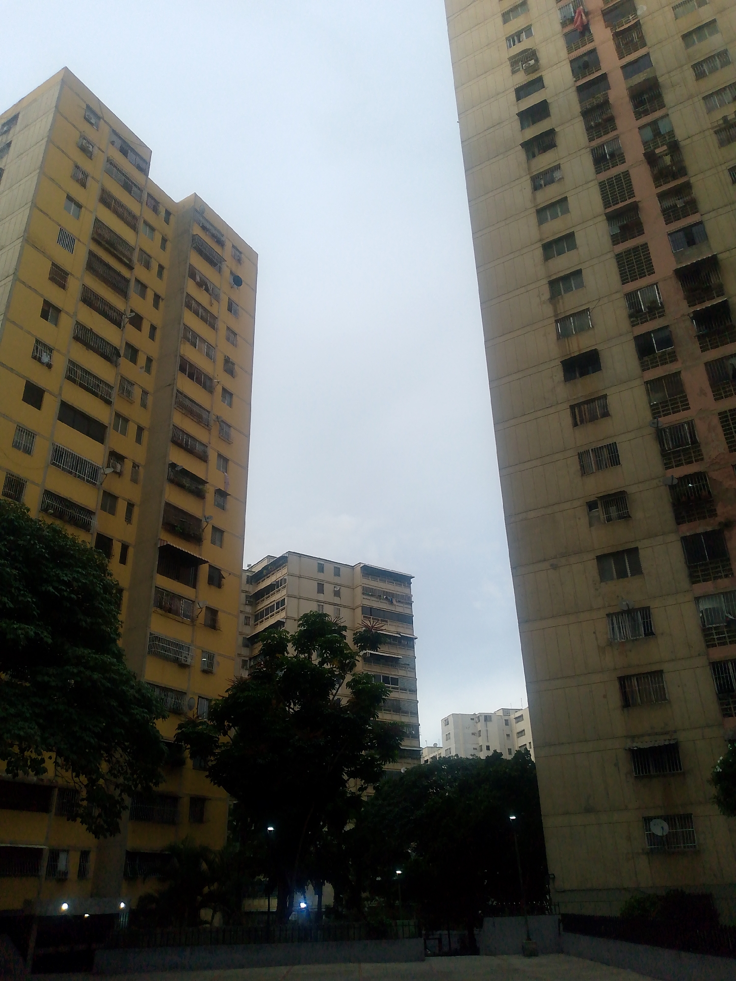 The image captures a view of several tall apartment buildings in a city setting, likely Caracas, Venezuela, given the architecture and style of the buildings. The sky is overcast, suggesting either early morning, late afternoon, or a cloudy day. The buildings are multi-story, with visible windows and balconies. The building on the left is predominantly yellow, while the building on the right is a lighter, neutral color, possibly beige or light brown, with pinkish accents on some sections. There are trees in the foreground and middle ground, some of which are dense and dark green, obscuring parts of the lower floors of the buildings. Several streetlights are visible in the darker, lower-left portion of the image, illuminating the ground level. The overall impression is one of an urban residential area, with a somewhat somber or muted atmosphere due to the overcast sky and the presence of shadows from the trees. There are no people or identifiable vehicles visible in the frame. The scene appears static and devoid of any immediate activity.
