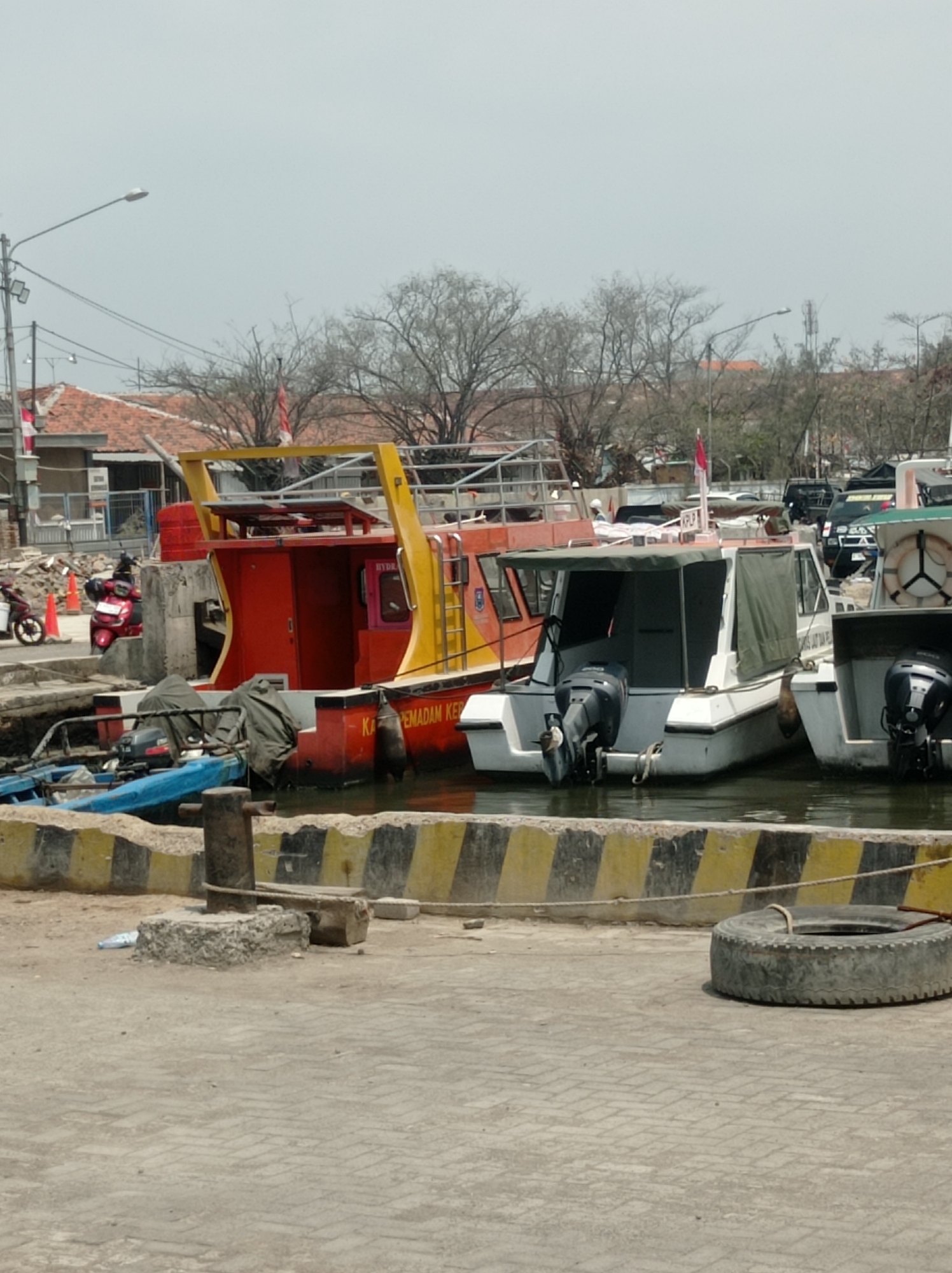 The image shows a harbor scene in Tegal, Indonesia, on a bright, likely midday, day. The sky is overcast, suggesting a hazy or cloudy atmosphere. The primary subjects are several boats docked at the harbor.
In the foreground, a paved area transitions to a concrete embankment with a yellow and black striped barrier. A large tire is resting on the pavement to the right. A metal bollard stands on the embankment in the center.
Several boats are visible. The most prominent is a large red and yellow vessel, likely a ferry or transport boat, with a cabin and a superstructure. It has "KA PEMADAM KE" visible on its side, suggesting it may be related to fire fighting or rescue. Next to it are two smaller white boats, each with an outboard motor. The boat on the right has a lifebuoy mounted on its side. A blue tarp covers part of a small boat or floating object in the water.
In the background, there are buildings with red-tiled roofs, trees with bare branches, and utility poles with streetlights. The presence of Indonesian flags suggests a patriotic or official setting. Red scooters are parked near the red and yellow boat, indicating activity and potential visitors. The overall impression is one of a functional working harbor with multiple vessels.