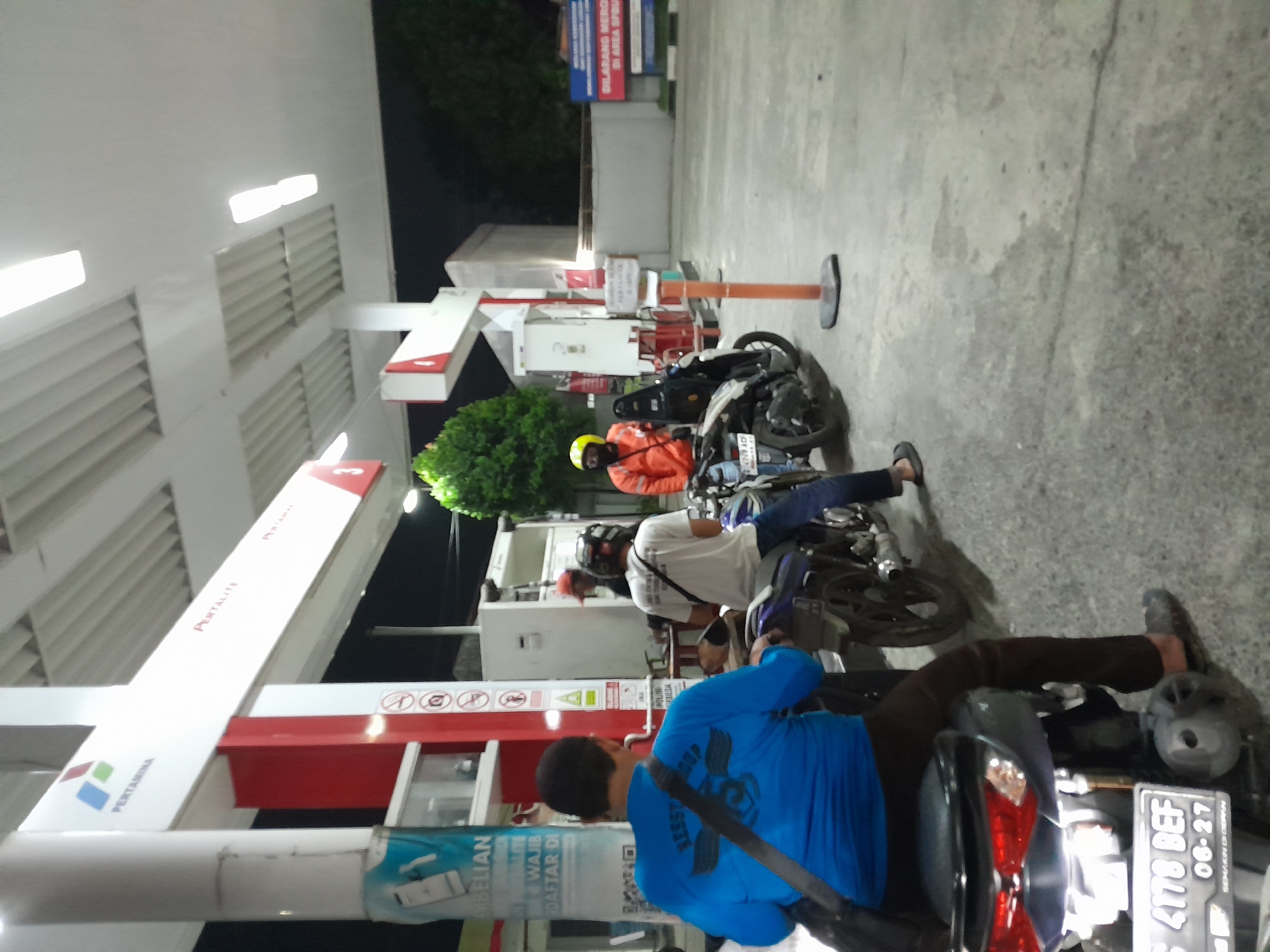 The image depicts a night scene at a Pertamina gas station in Kademangan, Indonesia. The gas station has a white canopy with integrated lighting, supported by white pillars. Red and white signage is visible on the structure, including the Pertamina logo. Several individuals on motorcycles are present. In the foreground, a person wearing a blue long-sleeved shirt and dark pants is seated on a motorcycle. To their right, another person in a white t-shirt and dark pants is also on a motorcycle. Further back, a person in an orange jacket and yellow helmet is on a motorcycle. In the background, there are trees and a building with a fence. A sign with red lettering that translates to "PROHIBITED TO SMOKE IN THE AREA" is visible. The ground is paved concrete. The overall atmosphere suggests a routine stop at a gas station during the evening.