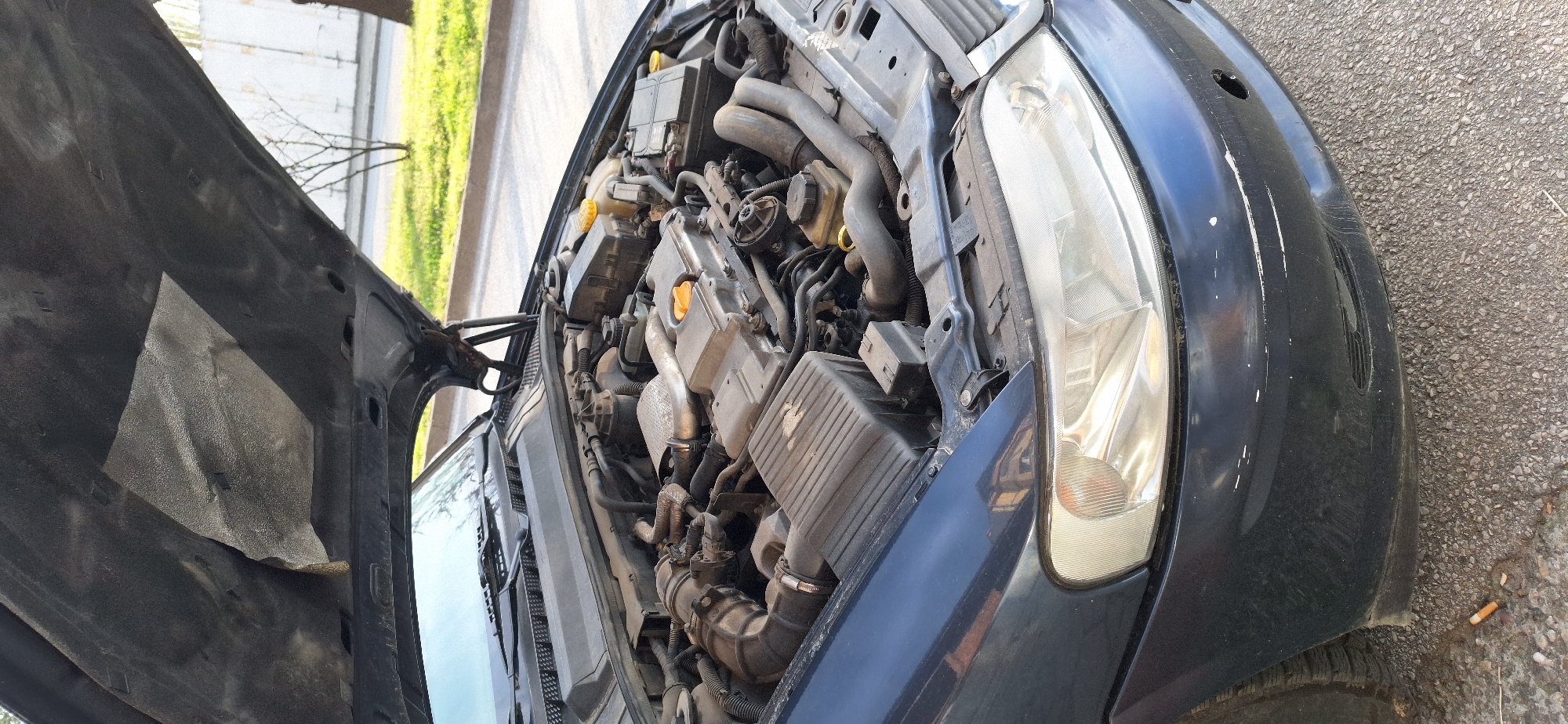 The image shows the open hood of a dark-colored car, revealing the engine compartment. The car is parked outdoors on an asphalt surface. The engine appears to be a standard internal combustion engine with various components such as hoses, manifolds, and boxes. The front of the car shows a headlight and part of the front fender with some visible scratches. The environment appears to be urban, possibly a street or parking lot, as greenery and buildings are visible in the background. The image does not provide specific information to identify the location in Blagoevgrad, Bulgaria.