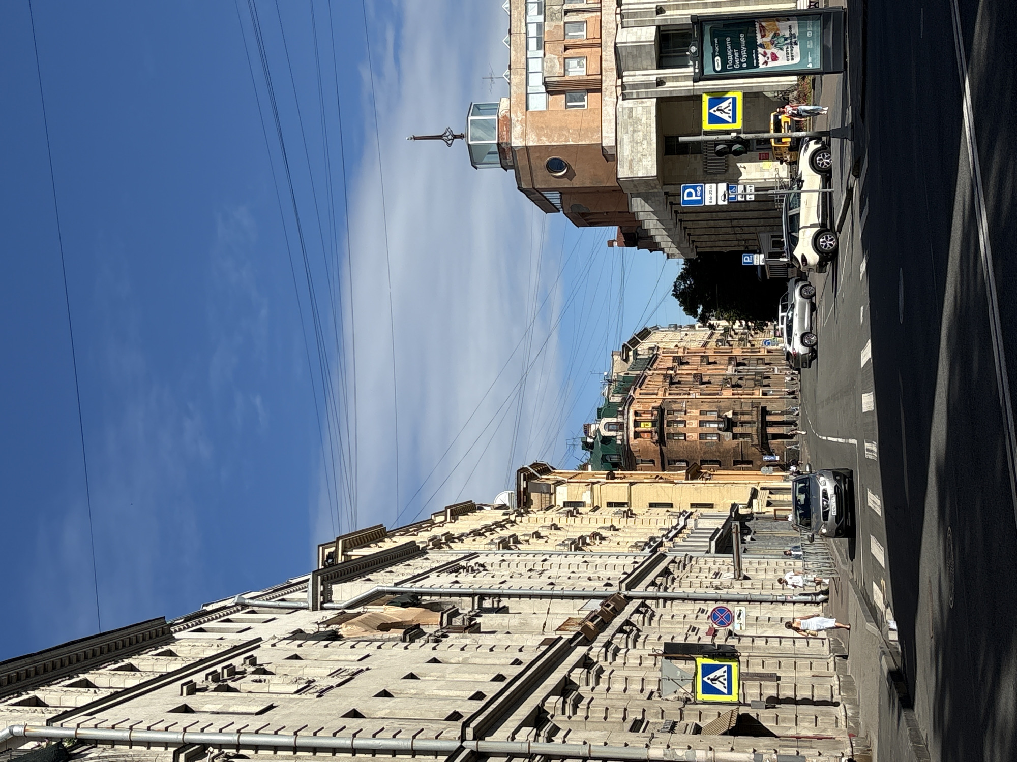 An eye-level, vertical, full shot captures a street scene in Saint Petersburg, Russia, under a partly cloudy sky with visible overhead wires. On the left, a multi-story building with intricate architectural details and many windows dominates the foreground. A pedestrian crossing sign with a blue and yellow pedestrian symbol is mounted on the building's facade. A woman in a white dress walks on the sidewalk, and a silver sedan is driving on the street.
The street stretches into the distance, lined with several other buildings of varying architectural styles and colors, including a distinctive orange building with a glass-topped turret on the right. Several cars are parked and moving on the road, and other pedestrians can be seen walking on the sidewalks. Traffic lights and street signs are visible, including a blue parking sign with times listed. A large advertisement poster is also present on the right side of the image. The overall impression is a busy urban street on a sunny day.