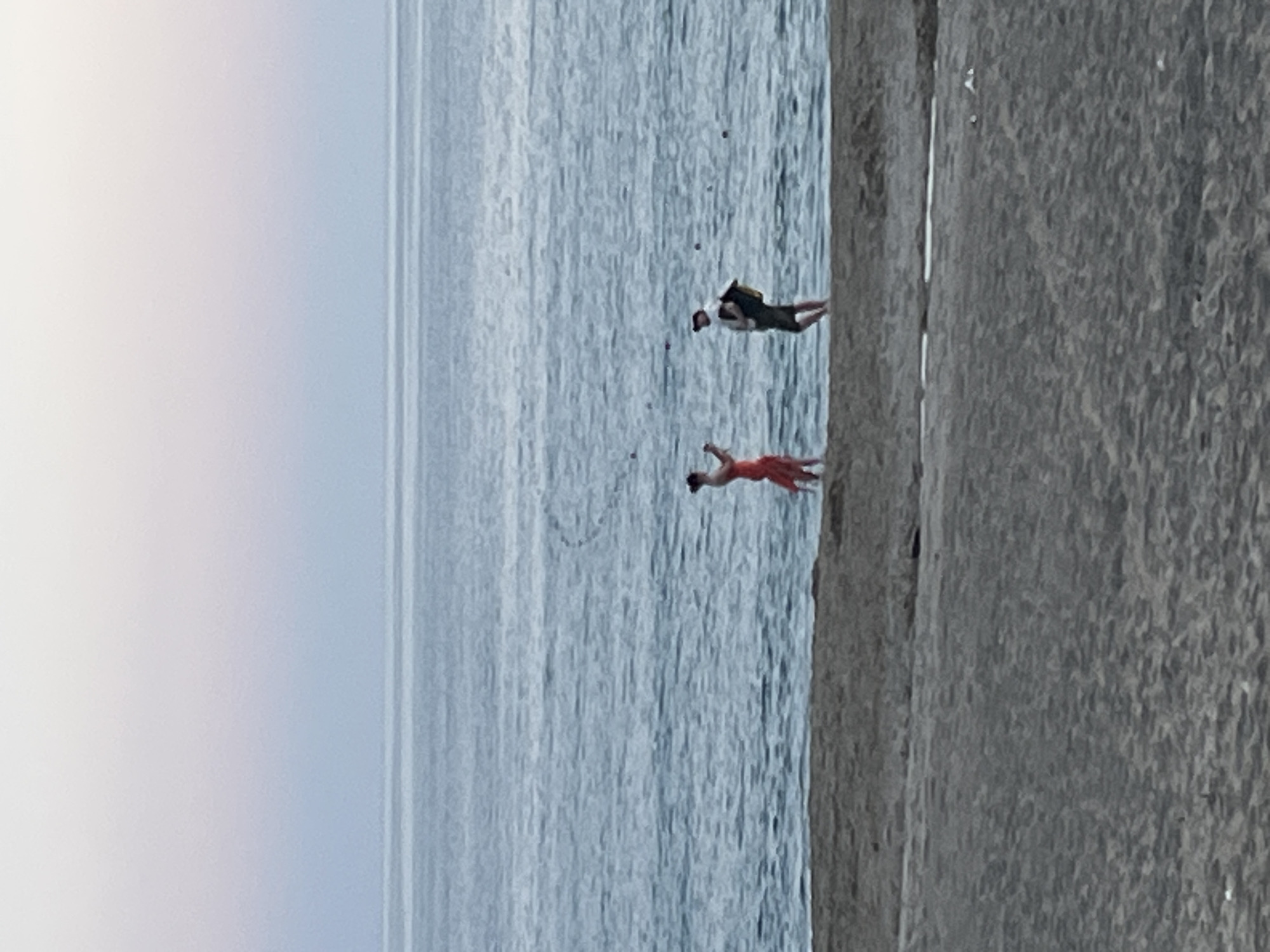 An image, correctly oriented, depicts a serene coastal scene in Alanya, Turkey. Two individuals are situated in the calm, blue-grey waters of the sea, near a sandy or rocky shoreline that occupies the right side of the frame. The sky, a soft, pale blue-grey, extends across the top, blending seamlessly into the horizon with the vast expanse of water.
The person further out in the water is wearing a dark top, dark shorts, and a backpack. They are partially submerged, appearing to be wading or standing in shallow water, facing away from the camera and towards the open sea. Closer to the shore, a second person is visible, clad in a striking red outfit, possibly a swimsuit or light summer wear. This individual also appears to be in the water, with their back mostly turned to the viewer, but their head is slightly angled. Their arms are raised and extended, suggesting movement, perhaps running into the water, swimming, or striking a dynamic pose.
The activity taking place appears to be recreational, with both individuals enjoying the sea. The water surface shows gentle ripples, indicating a calm day. The light is diffused and even, suggesting either an overcast sky or a time of day like early morning or late afternoon, without direct harsh sunlight. The overall atmosphere conveys a sense of peaceful leisure. There is no visible text in the image.