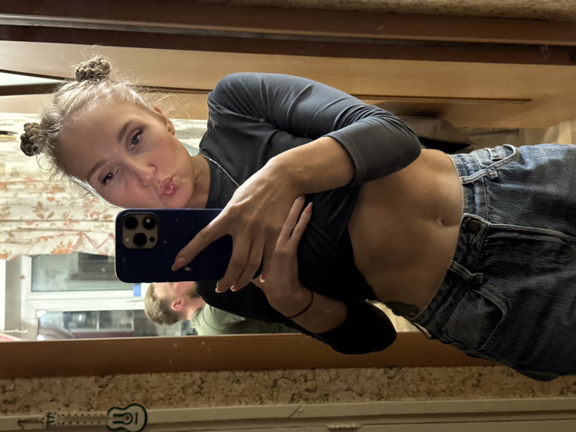 A woman with blonde hair styled in two buns is taking a selfie in front of a mirror. She is wearing a dark long-sleeved shirt and blue jeans, with a portion of her midriff exposed. She is holding a blue smartphone to take the picture. Reflected in the mirror is a man with short, light-colored hair and a green shirt. The reflection also shows a window and patterned wallpaper. A small guitar sticker is visible on the surface below the mirror. The location is unknown, as there is no indication of Perm, Russia.