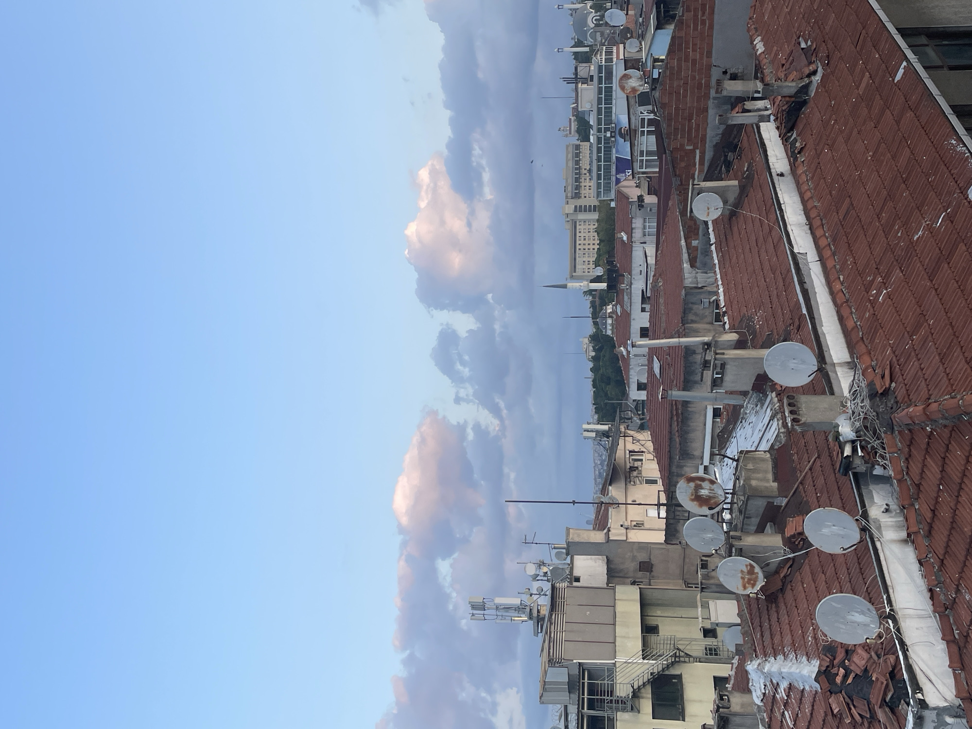 The image shows a rooftop view of a city, likely Kadıköy, Turkey, under a pale blue sky with scattered pink and white clouds. The foreground is dominated by red-tiled roofs, many of which appear weathered and have satellite dishes mounted on them. Several chimneys are also visible.
In the middle ground, there are buildings of varying heights and architectural styles. Some have light-colored facades, while others are darker. Antennas and communication towers are prominent on some of these structures. Further in the background, a mosque with a slender minaret stands out, alongside larger, modern buildings, some with visible windows. The distant cityscape fades into a hazy horizon, suggesting a dense urban environment. The overall impression is of a residential and commercial area. There are no people visible in the image.