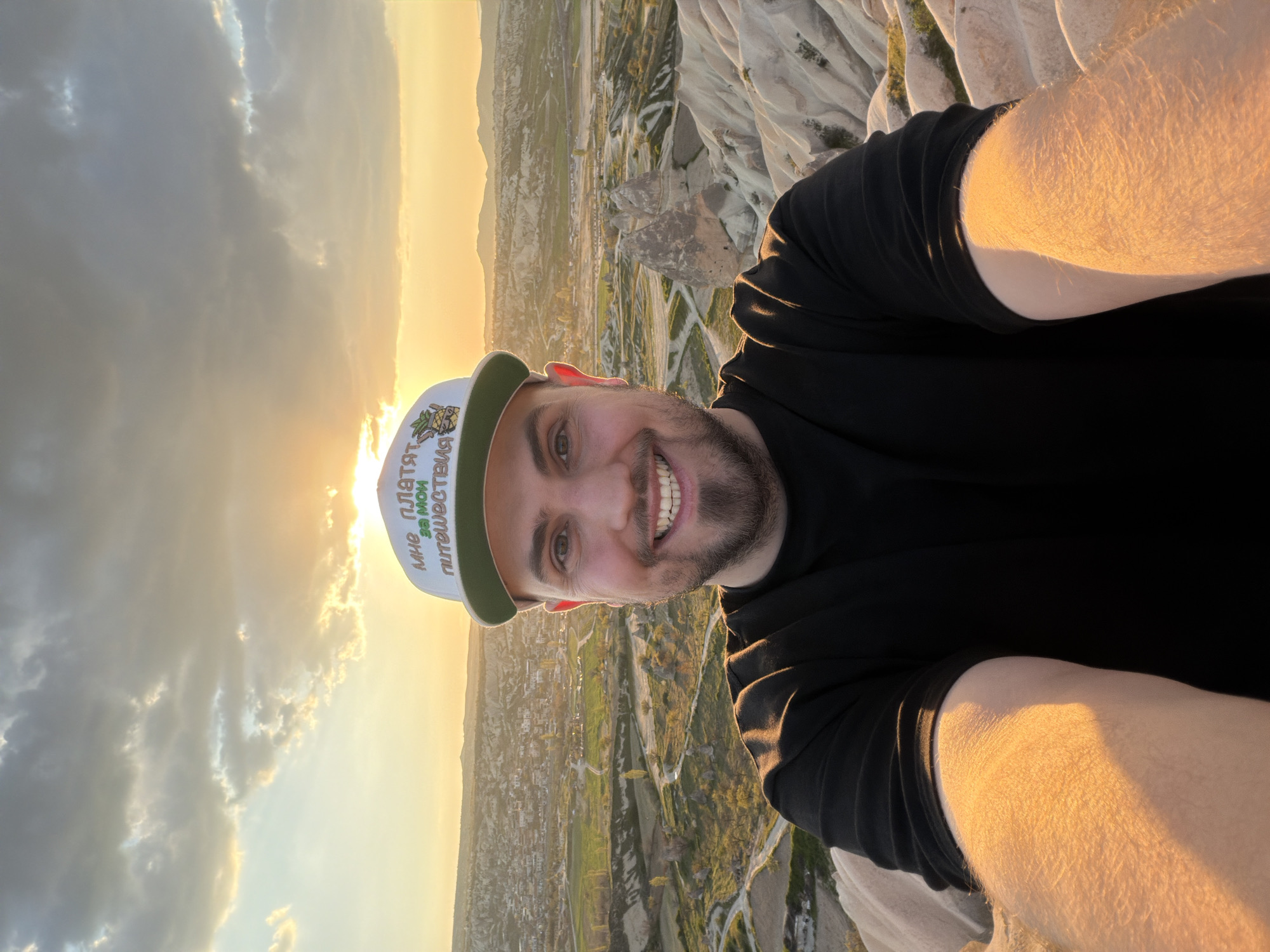 A man takes a selfie against a scenic backdrop at what appears to be sunrise or sunset, given the warm, low-angled light. The image is rotated 90 degrees counter-clockwise. The man is smiling broadly at the camera, with a beard and wearing a black t-shirt. He has a white baseball cap with a green brim. On the cap, text in Cyrillic script is visible, reading "МНЕ ПЛАТЯТ ЗА МОИ ПУТЕШЕСТВИЯ" which translates to "I AM PAID FOR MY TRAVELS," alongside a small graphical element resembling a pineapple.
The setting is an elevated vantage point, likely from a hot air balloon over Göreme, Turkey, as indicated by the distinctive "fairy chimney" rock formations and the panoramic view of the landscape. The background features rolling hills, white and light-colored rock formations, patches of greenery, and distant structures of a town or village. The sky shows scattered clouds, beautifully lit by the sun's golden rays, which also cast a warm glow on the man's skin and clothing. The scene conveys a sense of wonder and enjoyment.