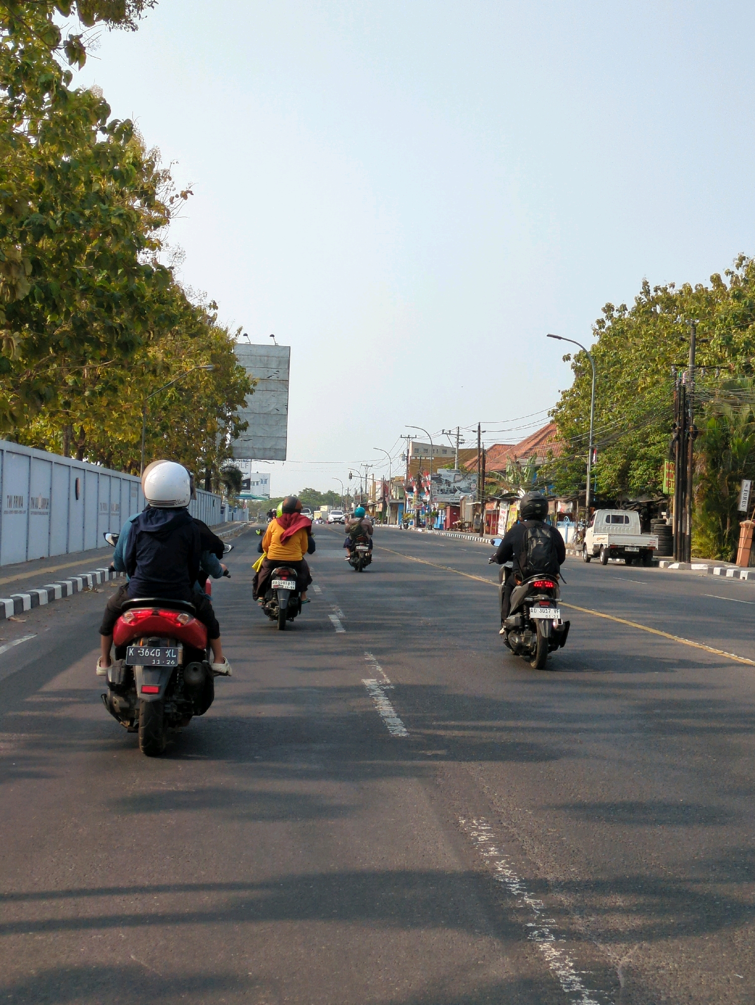 The image shows a view from a motorcycle traveling along a road in Colomadu, Indonesia. Several motorcycles are visible on the road, with riders wearing helmets and casual clothing. Trees and buildings line the sides of the road. The sky is clear and blue. The overall impression is that of a typical day in a busy Indonesian city.