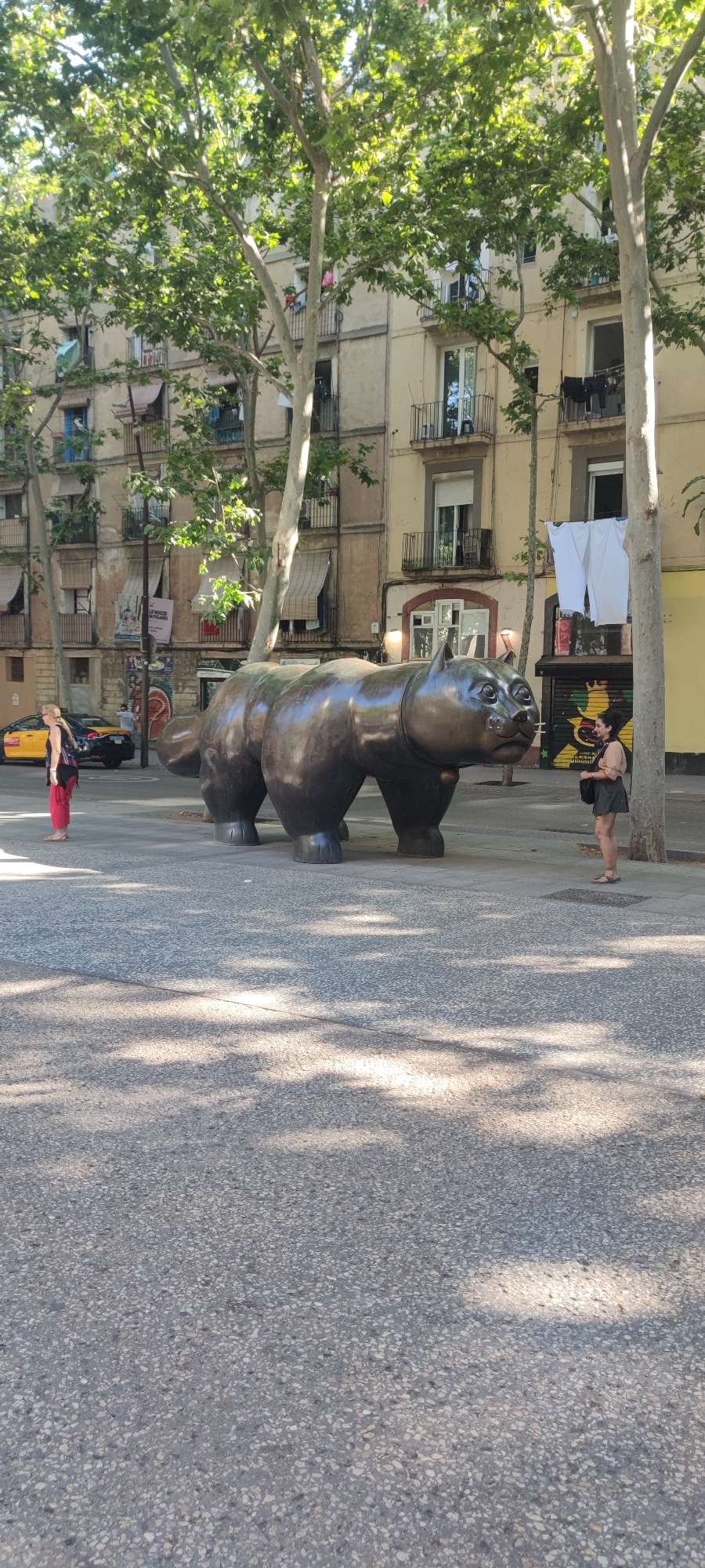 The image displays an urban scene in Barcelona, centered around a large, dark bronze sculpture of a plump cat, characteristic of Fernando Botero's "El Gato." The sculpture stands on light gray, speckled pavement, which is partially shaded by the canopy of numerous tall, leafy trees.
In the background, multi-story residential buildings with traditional European architecture line the street. These buildings feature multiple balconies, some equipped with awnings, and visible clotheslines where laundry, including white shirts and towels, hangs to dry.
Two women are present in the scene. On the left, a woman with blonde hair, wearing a black top and long red pants, is walking away from the viewer, barefoot, near a yellow taxi cab. On the right, a woman with dark hair, dressed in a light-colored top and dark skirt, stands facing the cat sculpture, observing it.
The setting is an outdoor plaza or wide street, bathed in bright daylight, indicated by the strong sunlight and distinct shadows cast by the trees. The time of day appears to be midday or early afternoon. A large banner or poster is partially visible on one of the buildings in the left background, but its text is illegible. The yellow taxi cab also has black text on its side. The atmosphere is calm, depicting an everyday street scene.