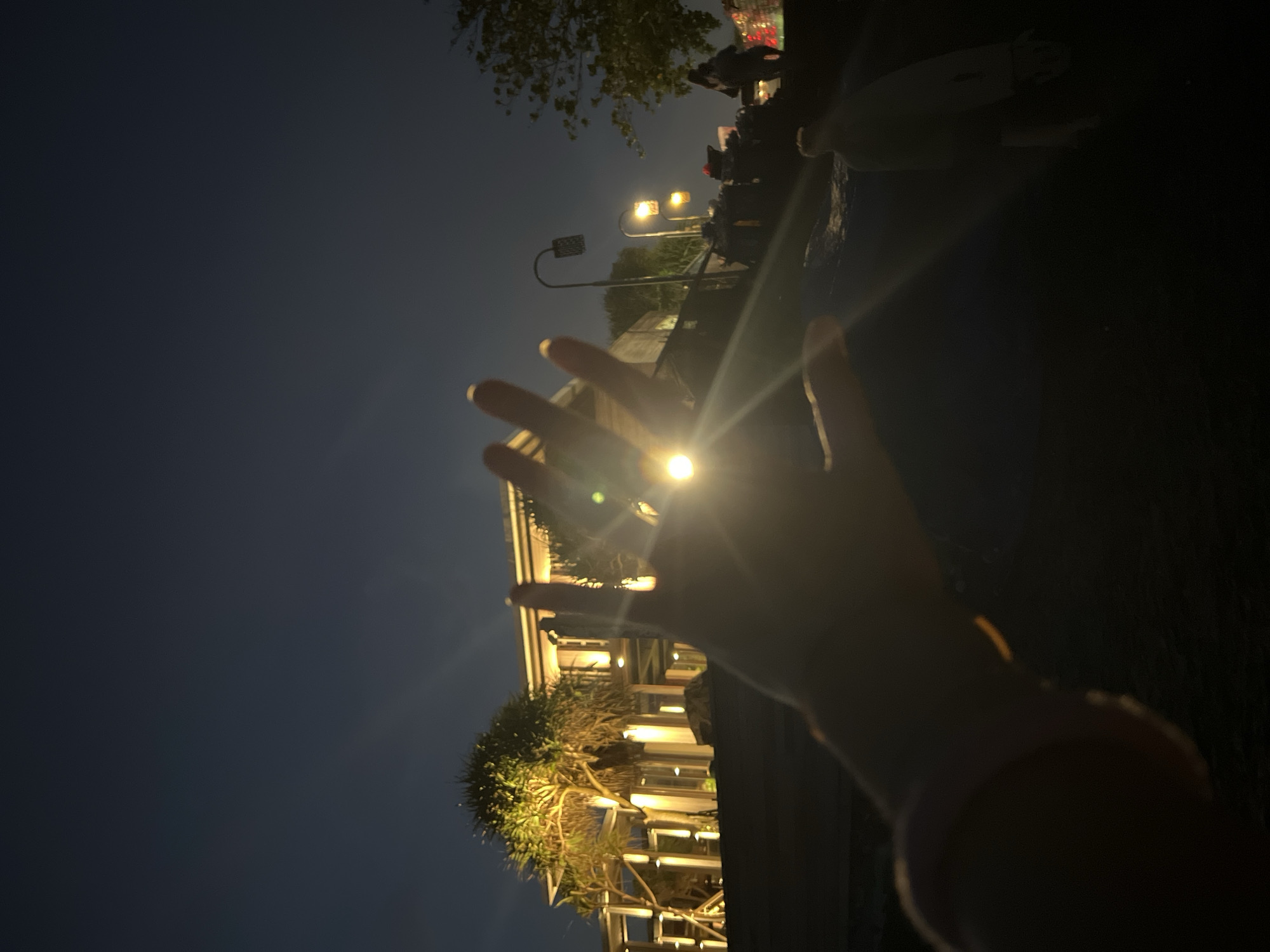 A low-angle, night-time photograph shows a hand obscuring a street lamp, taken in Canggu, Indonesia. The sky is a dark blue. In the foreground, a blurred hand is positioned in front of a bright street lamp, causing a lens flare. In the background, palm trees, a building with lights, and other street lamps are visible. The image is slightly rotated clockwise. A few people are barely visible on the right.